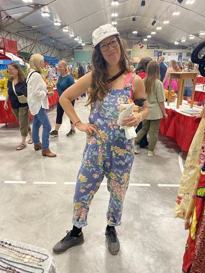 A woman in floral Butterflies in Ballgowns sustainably made and one of a kind overalls, gray sneakers, white cap, and glasses, posing at the Butterflies in Ballgowns market booth at the Rossland Fall Fair