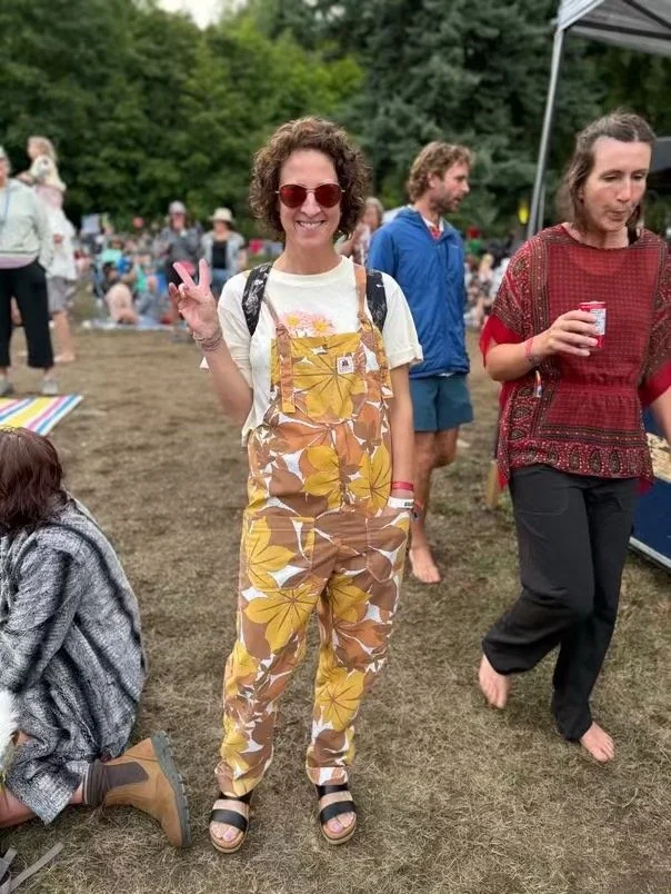 A woman smiling and making a peace sign with her hand at Kaslo Jazz Fest, wearing yellow leaf print Butterflies in Ballgowns overalls made sustainably in Nelson BC from an upcycled bed sheet.