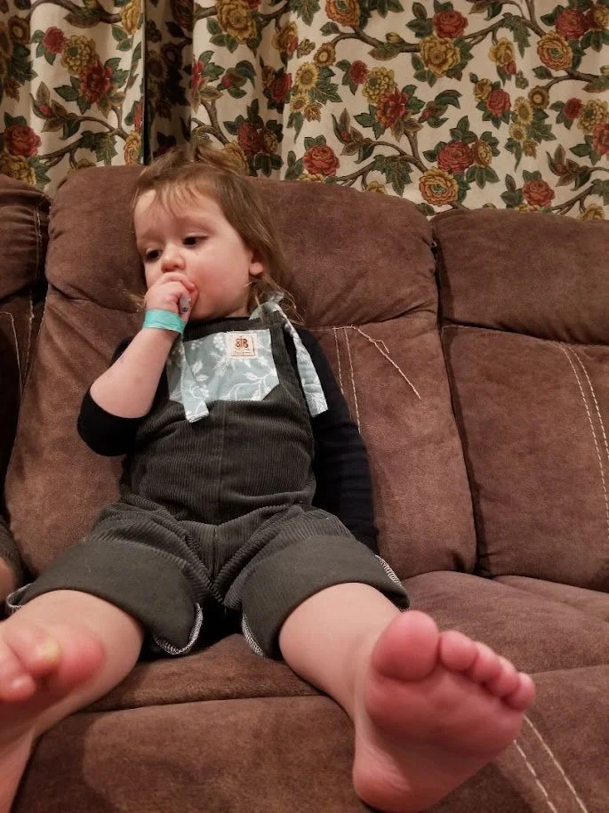 a male toddler, sat on a couch, wearing a custom made pair of butterflies in ballgowns one of a kind and sustainably made dungarees, in dark green corduroy with a blue floral pocket