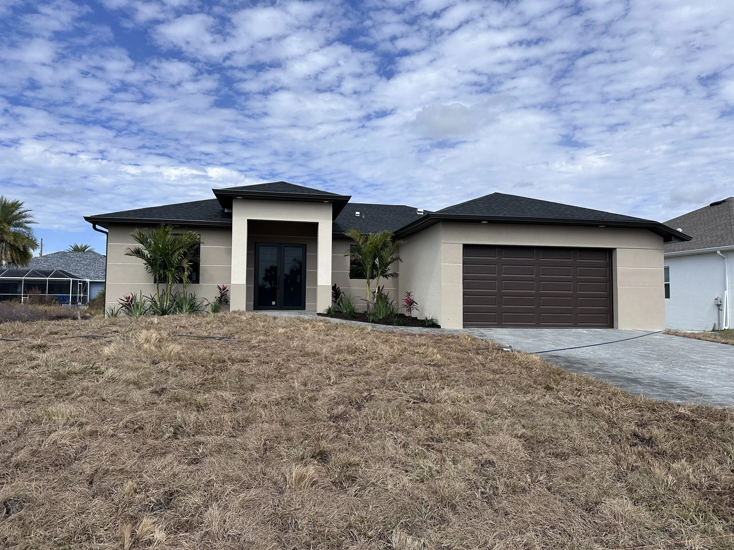 Single-story modern house with dark roof, beige walls, black front door, brown garage door, front garden with palms and plants, gray paving stone driveway, under partly cloudy sky.