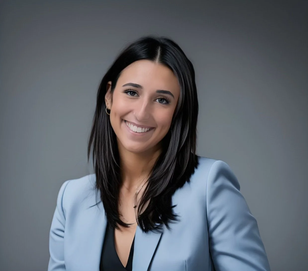 Portrait of a smiling woman with dark hair, wearing a light blue blazer and black top, against a gray background.
