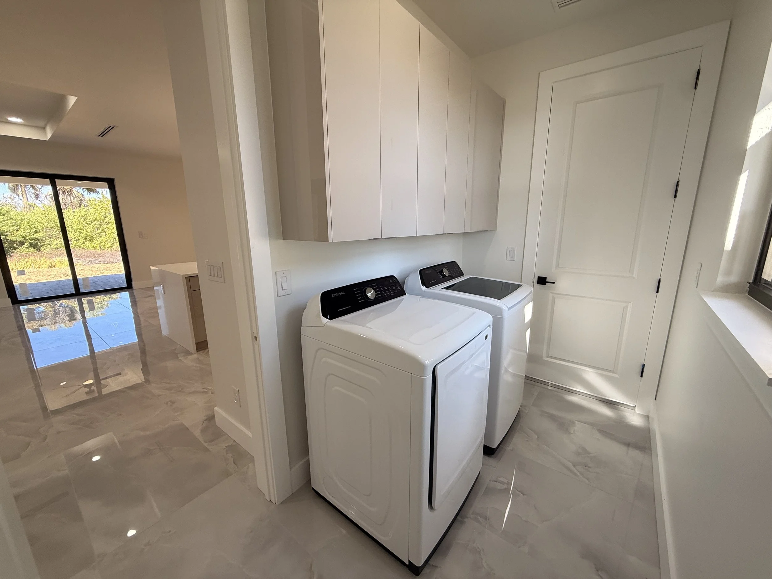Laundry room with a white washing machine and dryer side by side, beige cabinets above, white walls, and a window with sunlight.