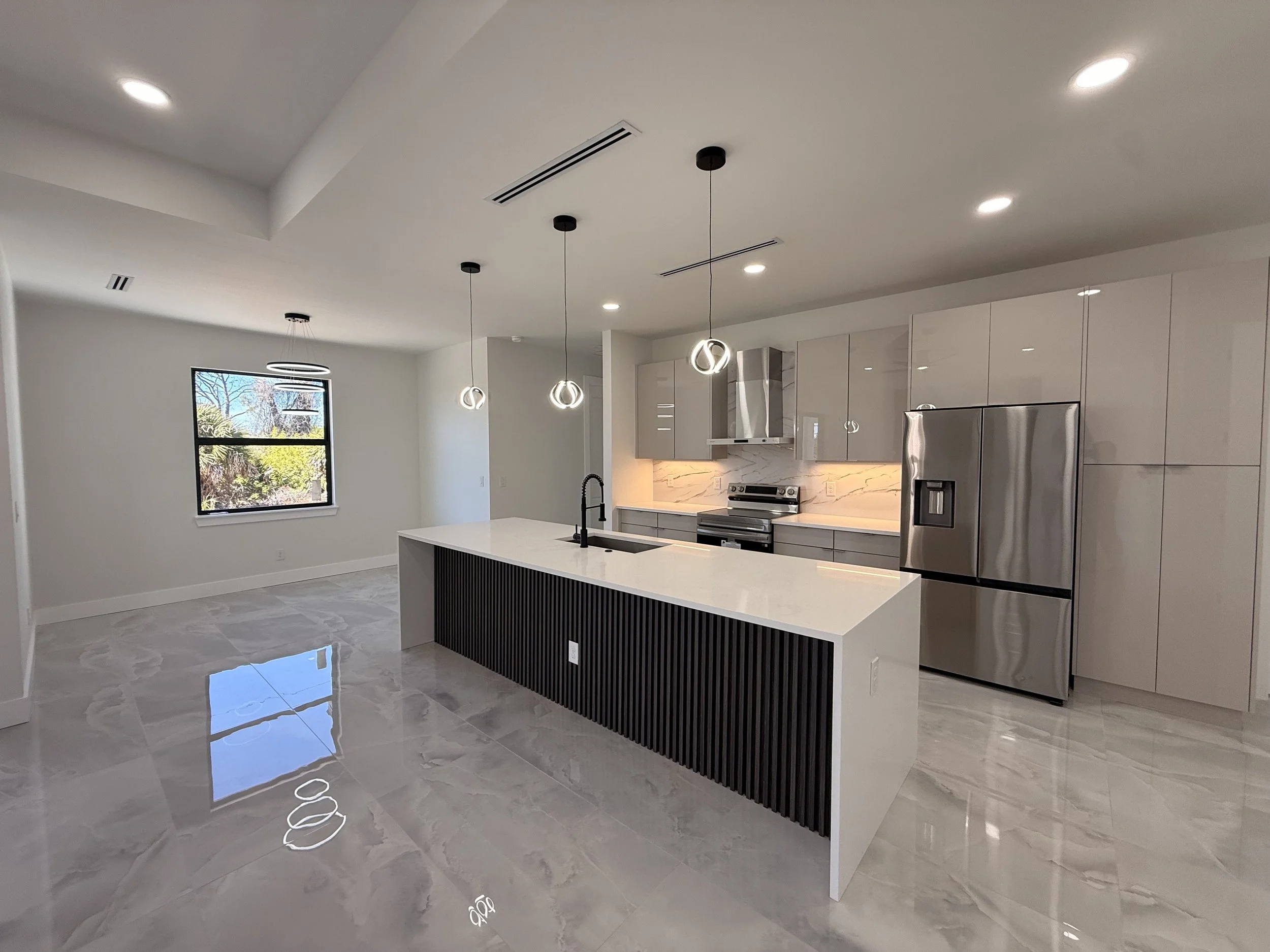 Modern kitchen with white walls, marble tile flooring, stainless steel appliances, a large kitchen island with a black slatted base, pendant lights, and a large window revealing trees outside.