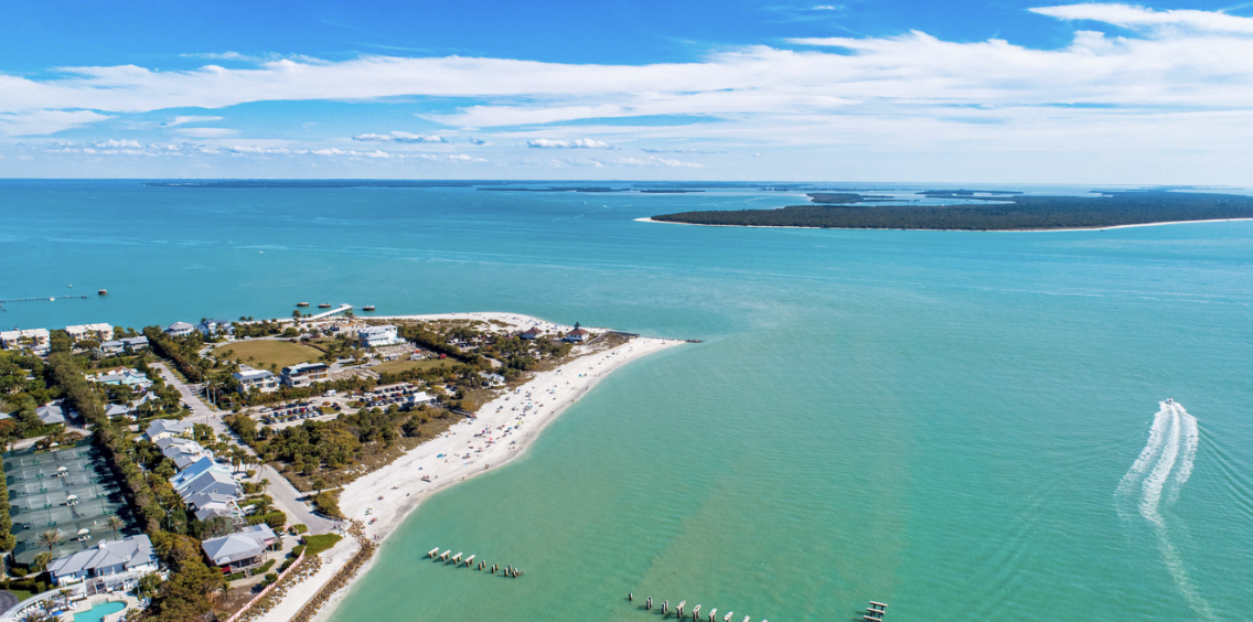 Aerial view of a coastal area with a white sandy beach, turquoise ocean, and oceanfront buildings surrounded by greenery. A boat creates wake on the water, and a distant island is visible on the horizon.