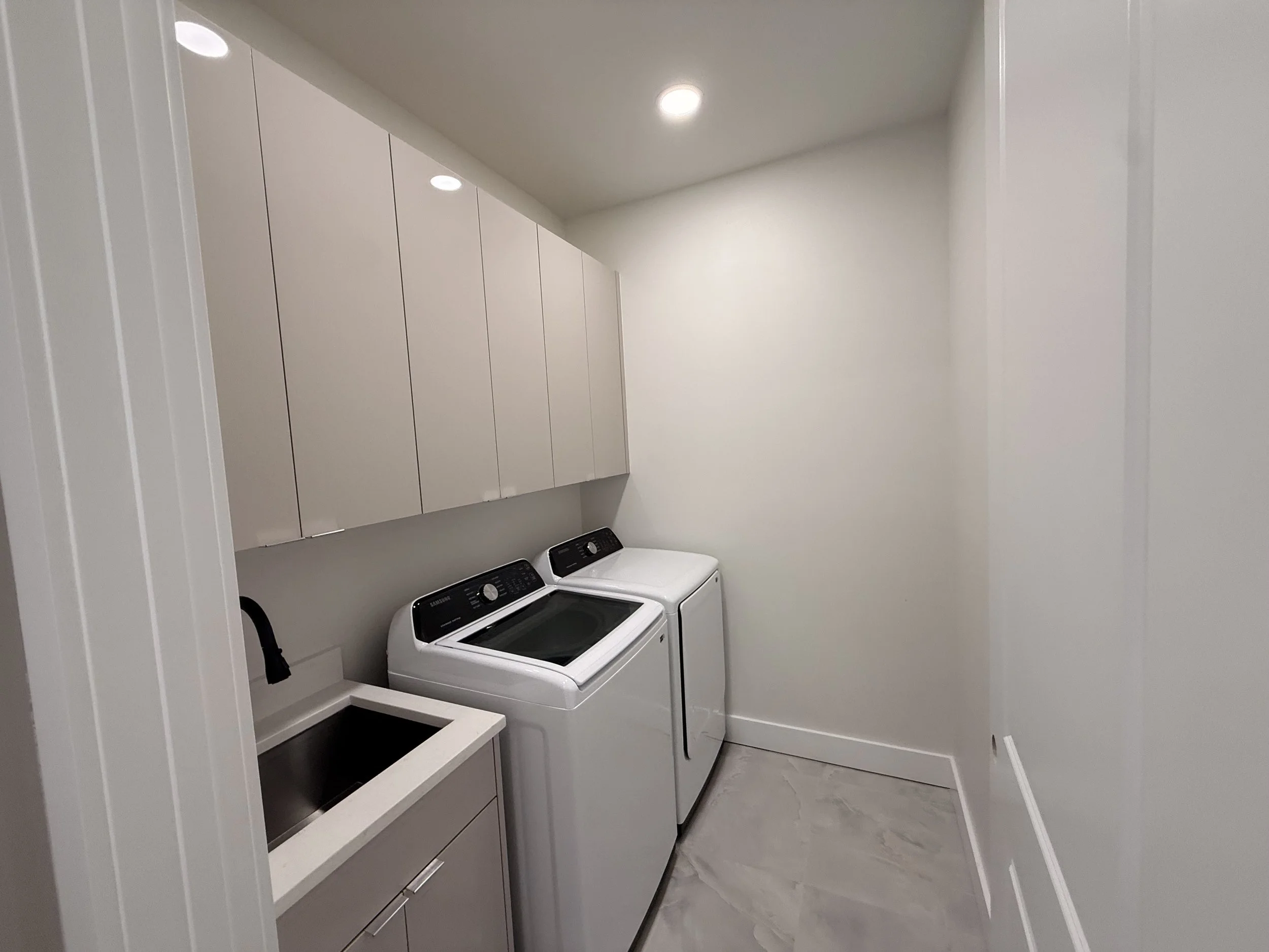 A modern laundry room with white walls, beige cabinets, a laundry sink, a washing machine, and a dryer.