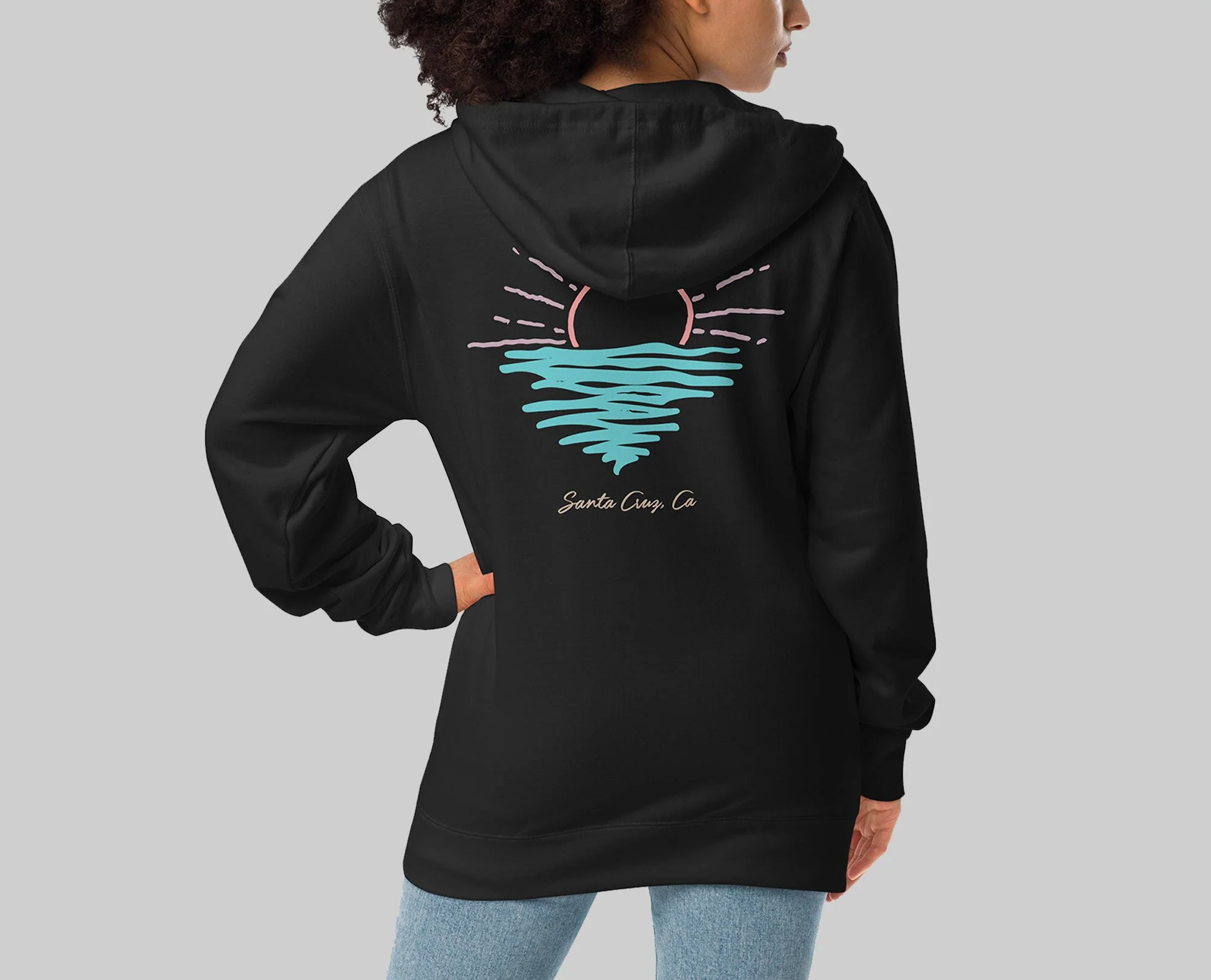 A woman wearing a black Santa Cruz Surf Teez hoodie with a pastel sunset and ocean wave graphic on the back, featuring pink sun rays, turquoise water, and the text “Santa Cruz, CA.”