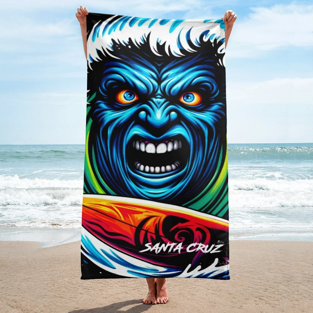 A person holding a colorful beach towel with a fierce blue wave man and the text 'Santa Cruz' on the beach on the bottom of the surfboard, with the ocean and sky in the background.