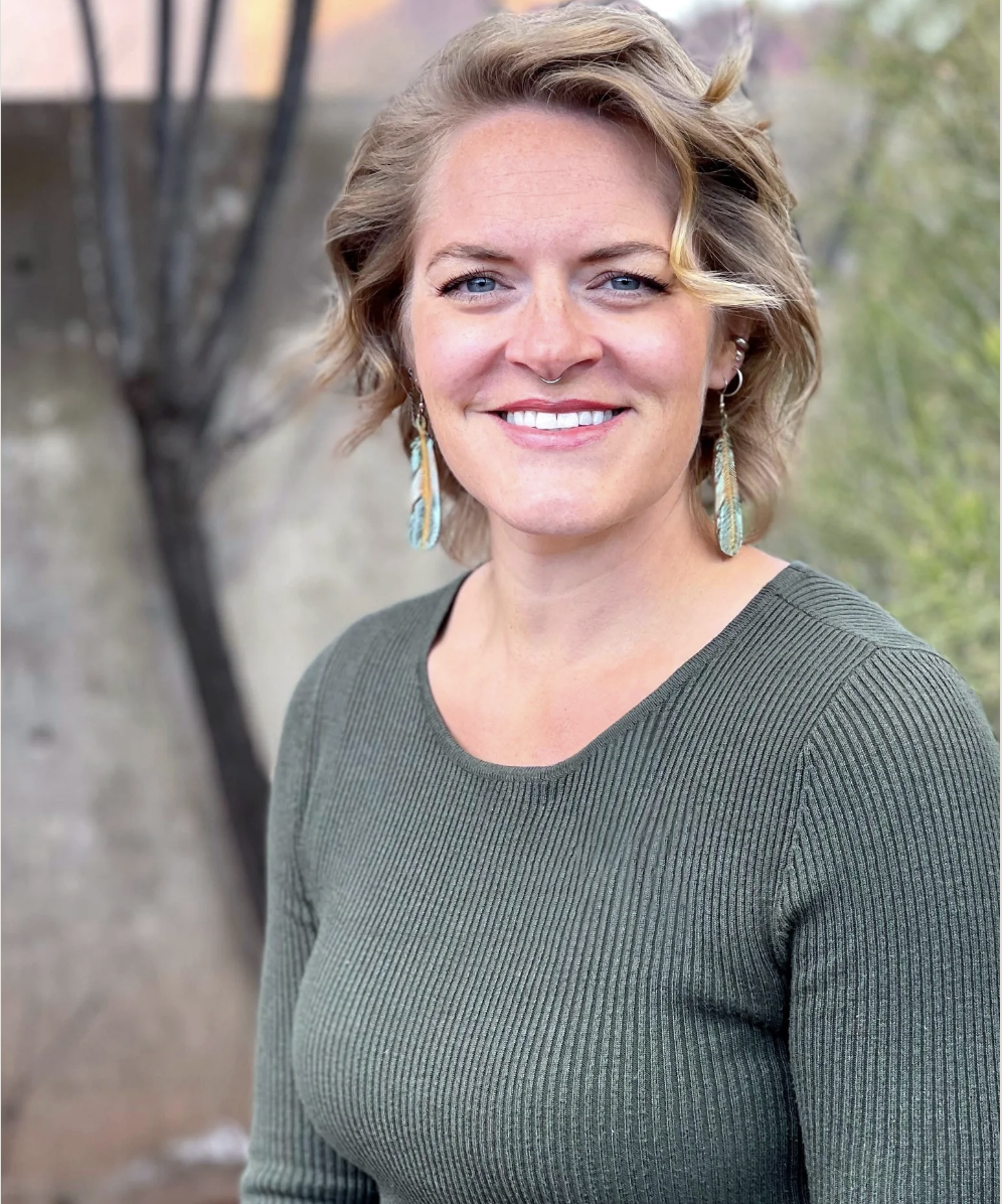 A woman with short wavy blonde hair and blue eyes, smiling, wearing a green ribbed long-sleeve shirt and feather earrings, standing outdoors with blurred background of trees and a concrete structure.
