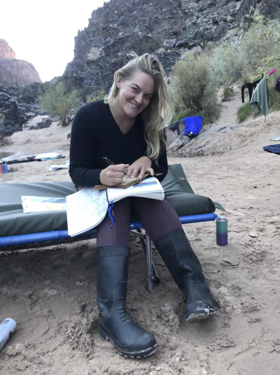 A woman sitting on a camping cot in a desert, wearing black boots, smiling, taking notes with a pen, with rocks, bushes, and a canyon in the background.