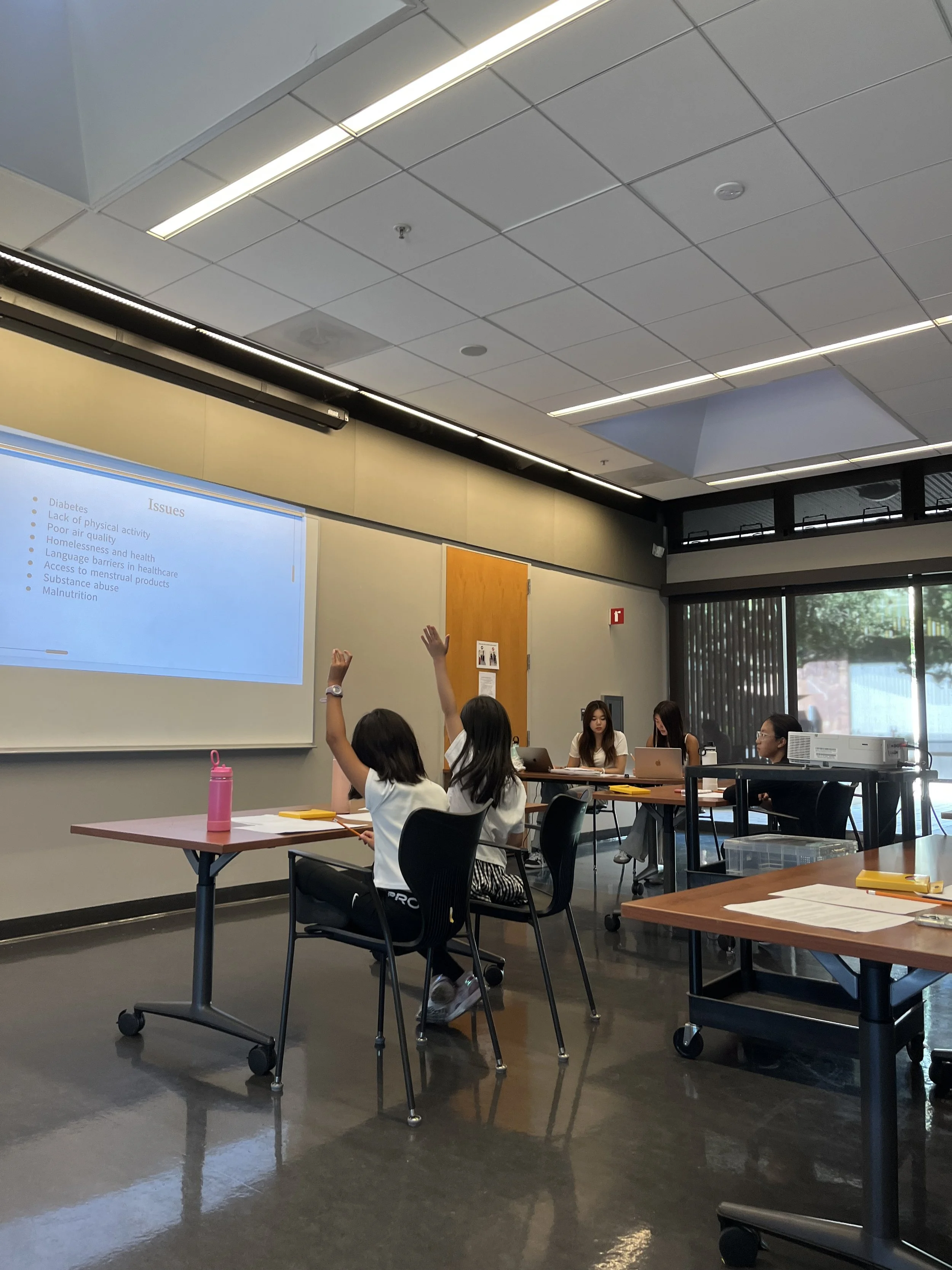 A classroom or meeting room with students raising their hands, two students seated at a table with a pink water bottle, and three students working on laptops at a different table. A projector is on a cart, and glass doors with curtains are in the background.