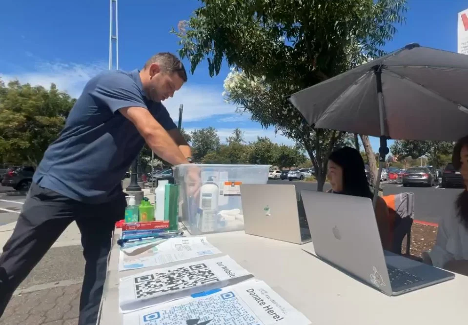 Man setting up a table with hand sanitizer, papers, and QR codes outdoors. Two women using laptops under a large umbrella, with a parking lot and trees in the background.