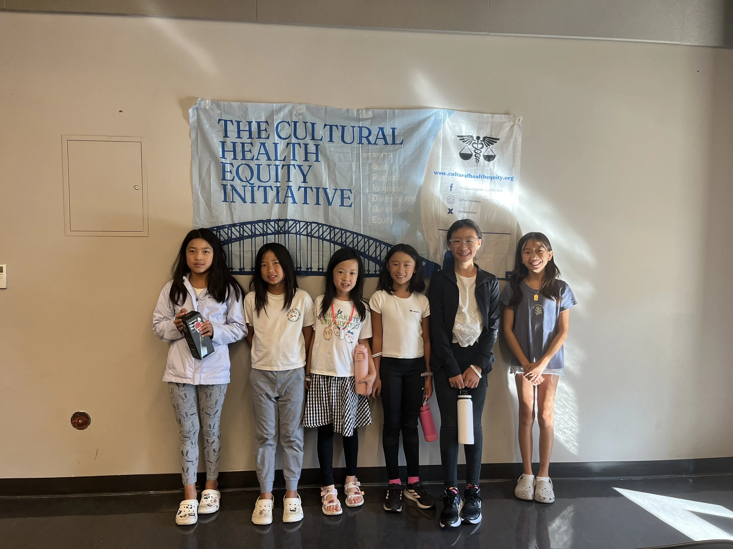 Six girls standing in front of a banner that reads 'The Cultural Health Equity Initiative' with a bridge illustration and social media icons.
