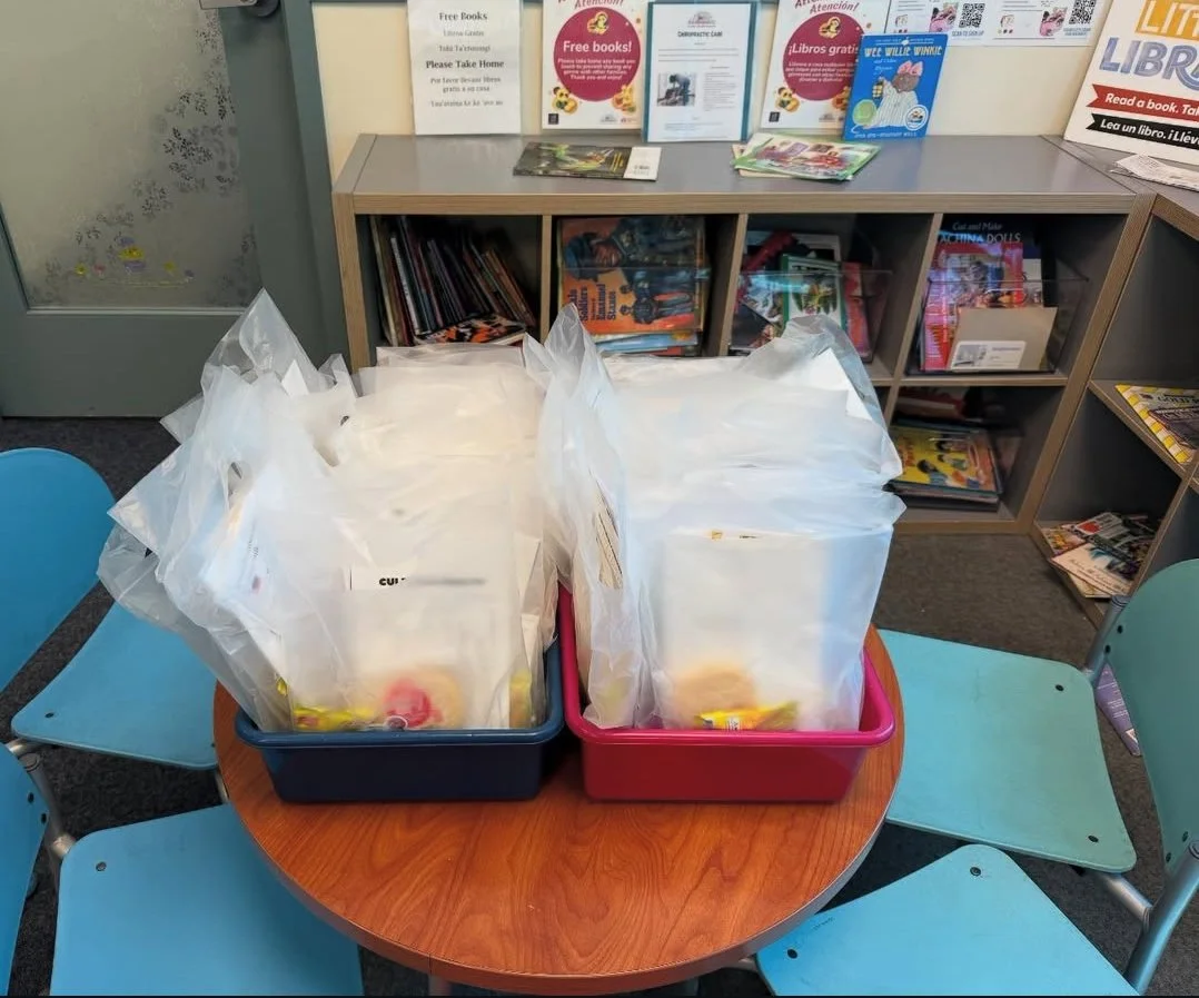 Two plastic bins filled with packaged items on a round wooden table in a library section with bookshelves in the background.