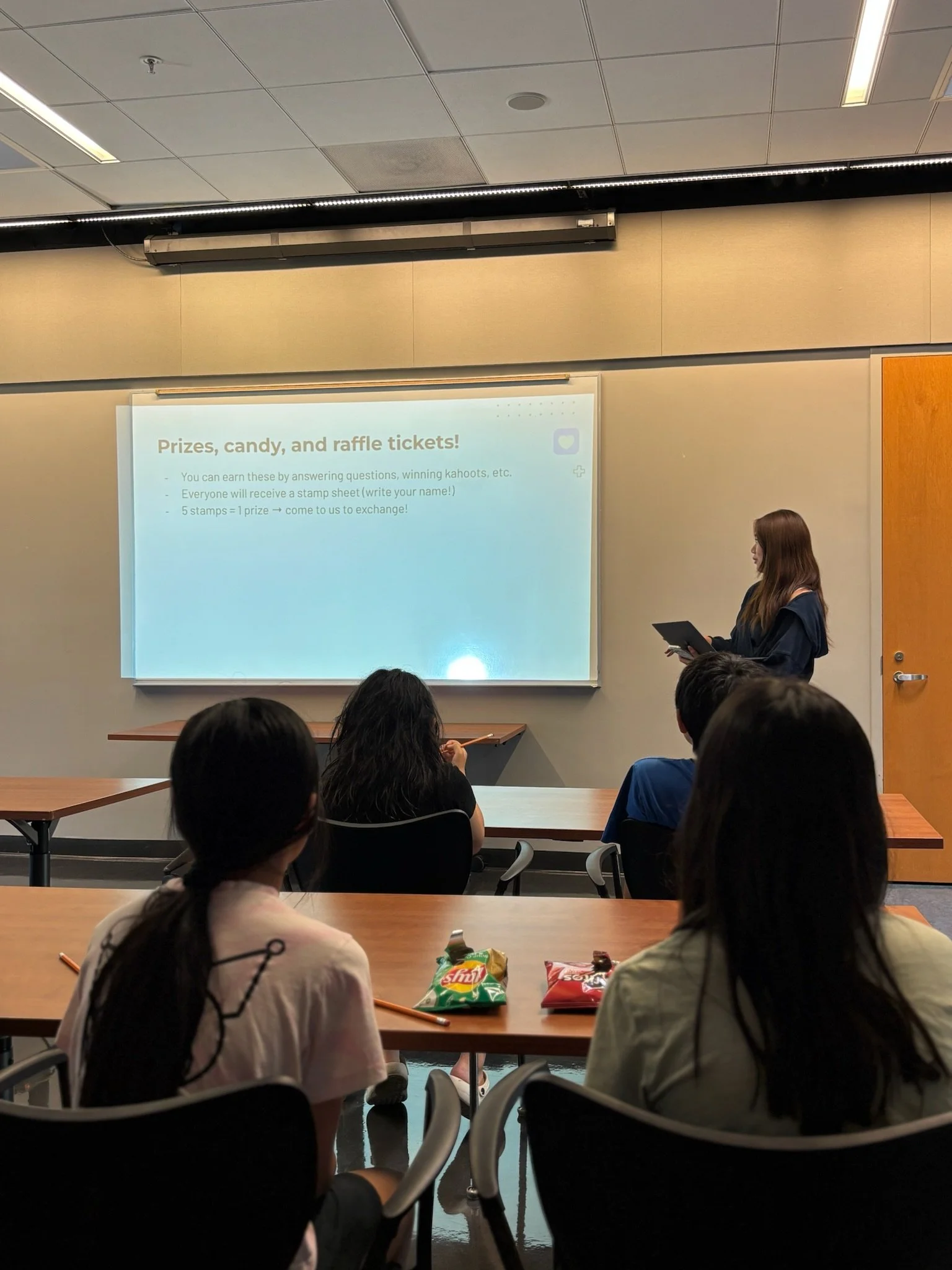 A classroom presentation about prizes, candy, and raffle tickets. A woman is standing at the front holding a tablet, and five people are sitting at tables looking at the presenter. There are bags of snacks on the tables, and a projector screen displaying information about earning raffle tickets.