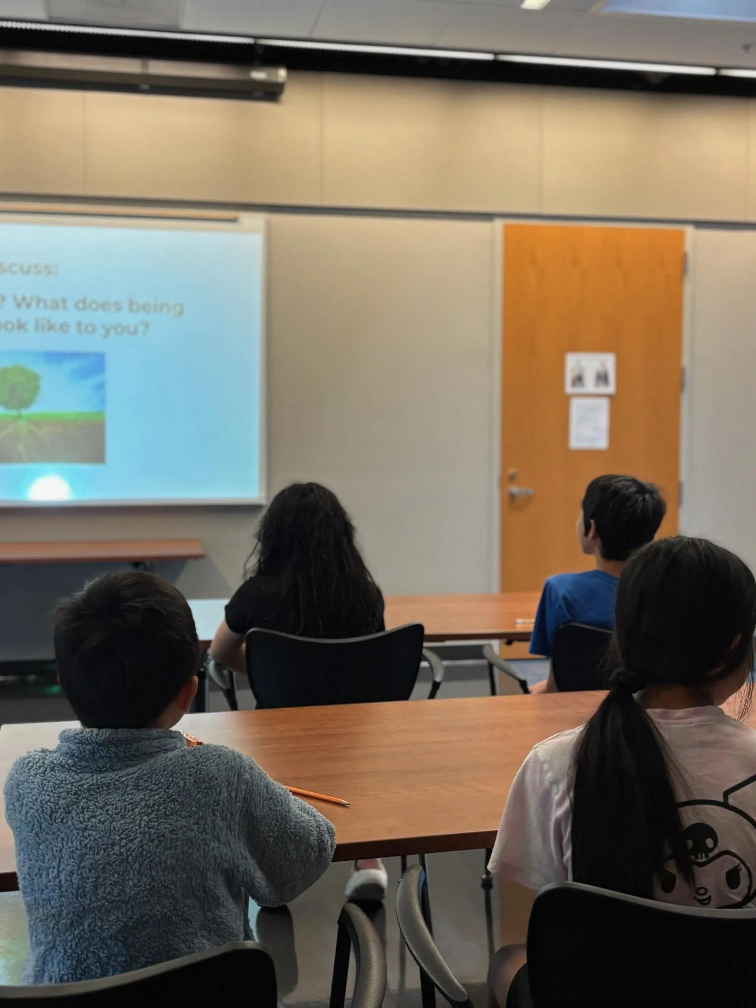 A classroom with four children sitting at desks, facing a projected presentation on the front wall. The presentation slide is partially visible and asks, 'What does being kind look like to you?' The children are focused on the screen.