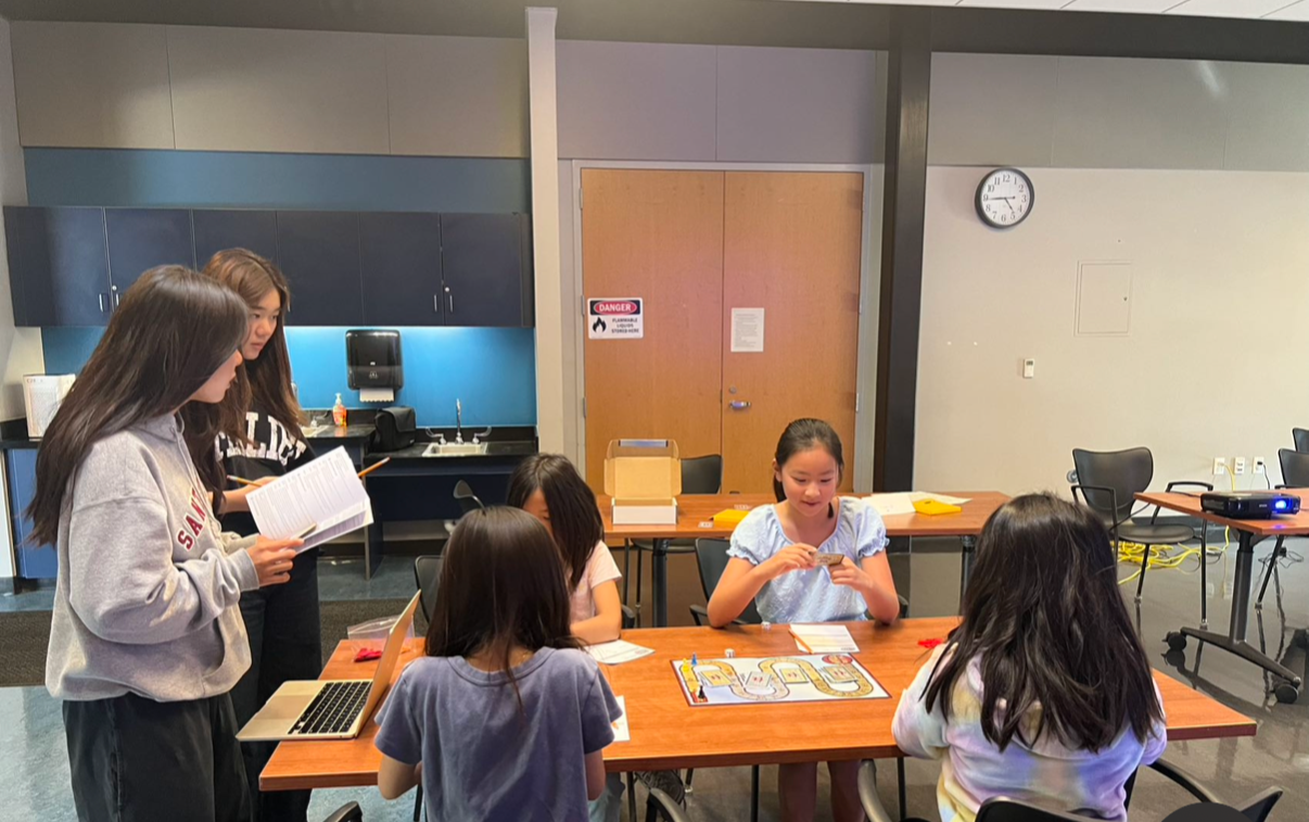 Five children in a classroom engaged in an activity with papers and a board game on a table, with two girls standing and three sitting.