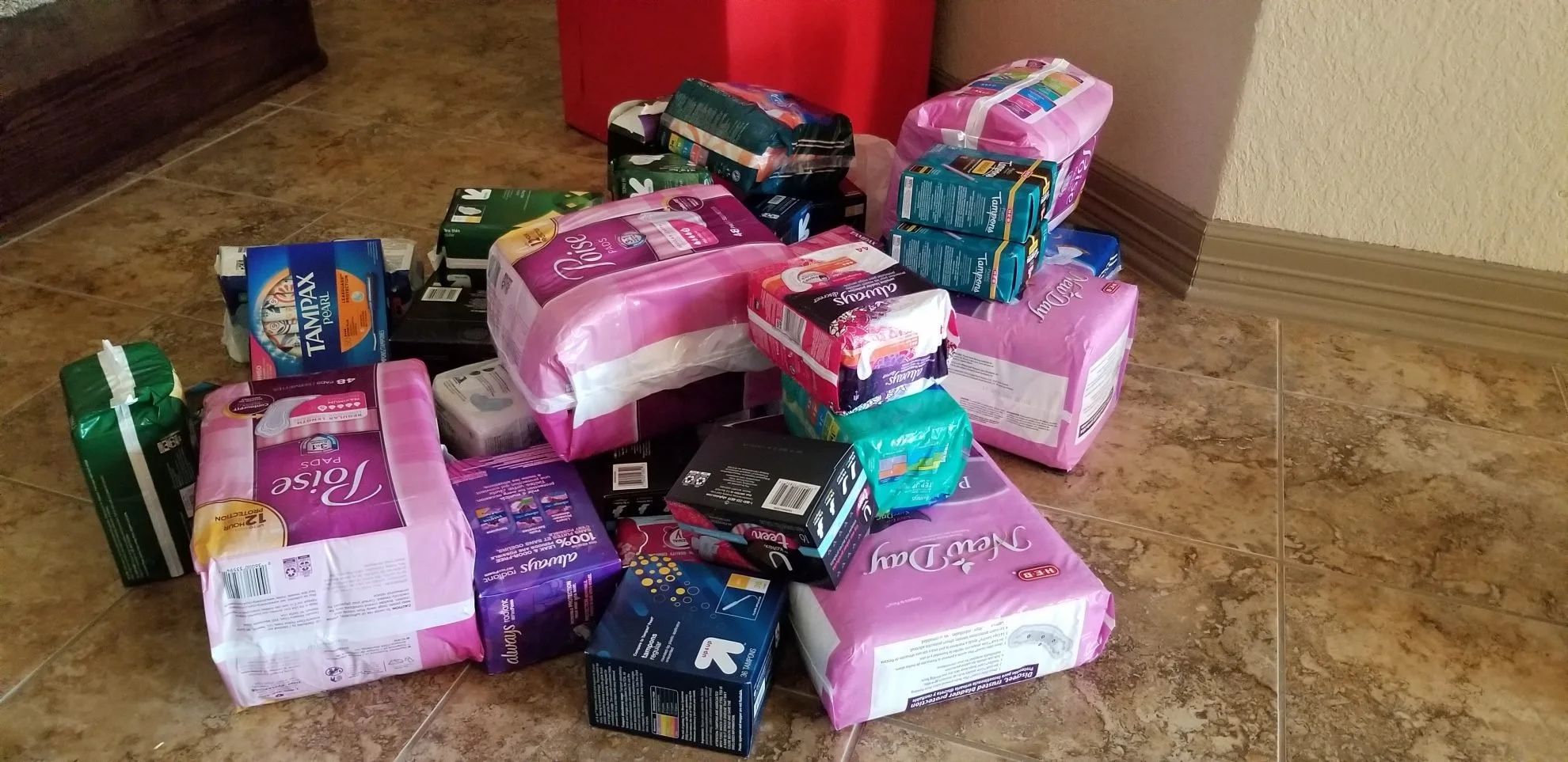 Piles of various brands and sizes of adult diapers, including packages of Depend, Tampax Pearl, and others, on a tiled floor near a wall corner.