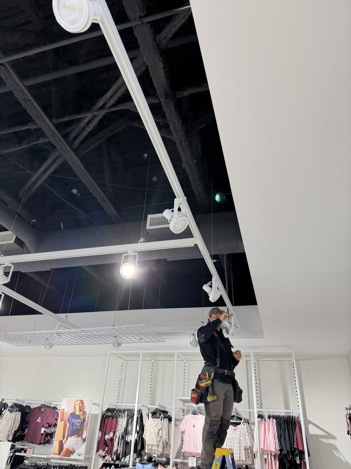 Koop Electrical electrician on a ladder replacing a track lighting head above the fitting rooms at Ardene, Southcentre Mall, Calgary, with the store's open black ceiling behind the white track