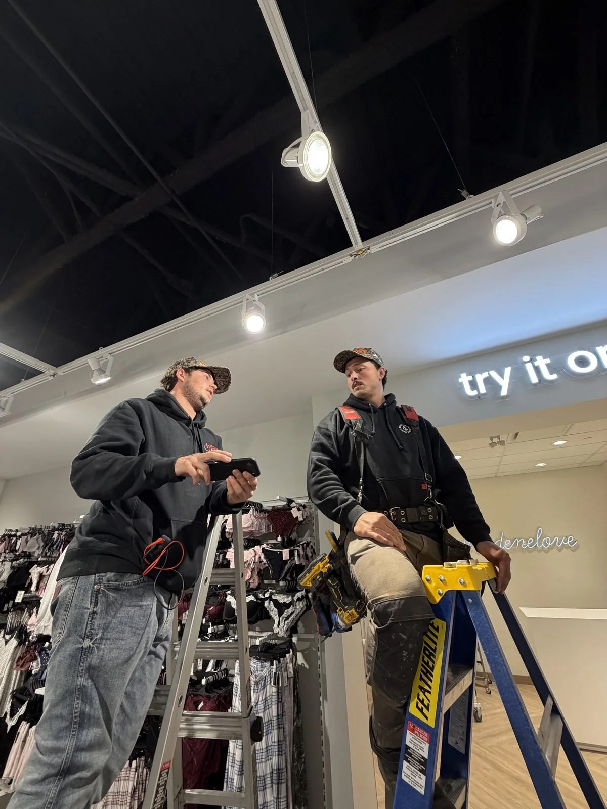 Two Koop Electrical electricians on ladders under newly lit track heads at Ardene, Southcentre Mall, Calgary