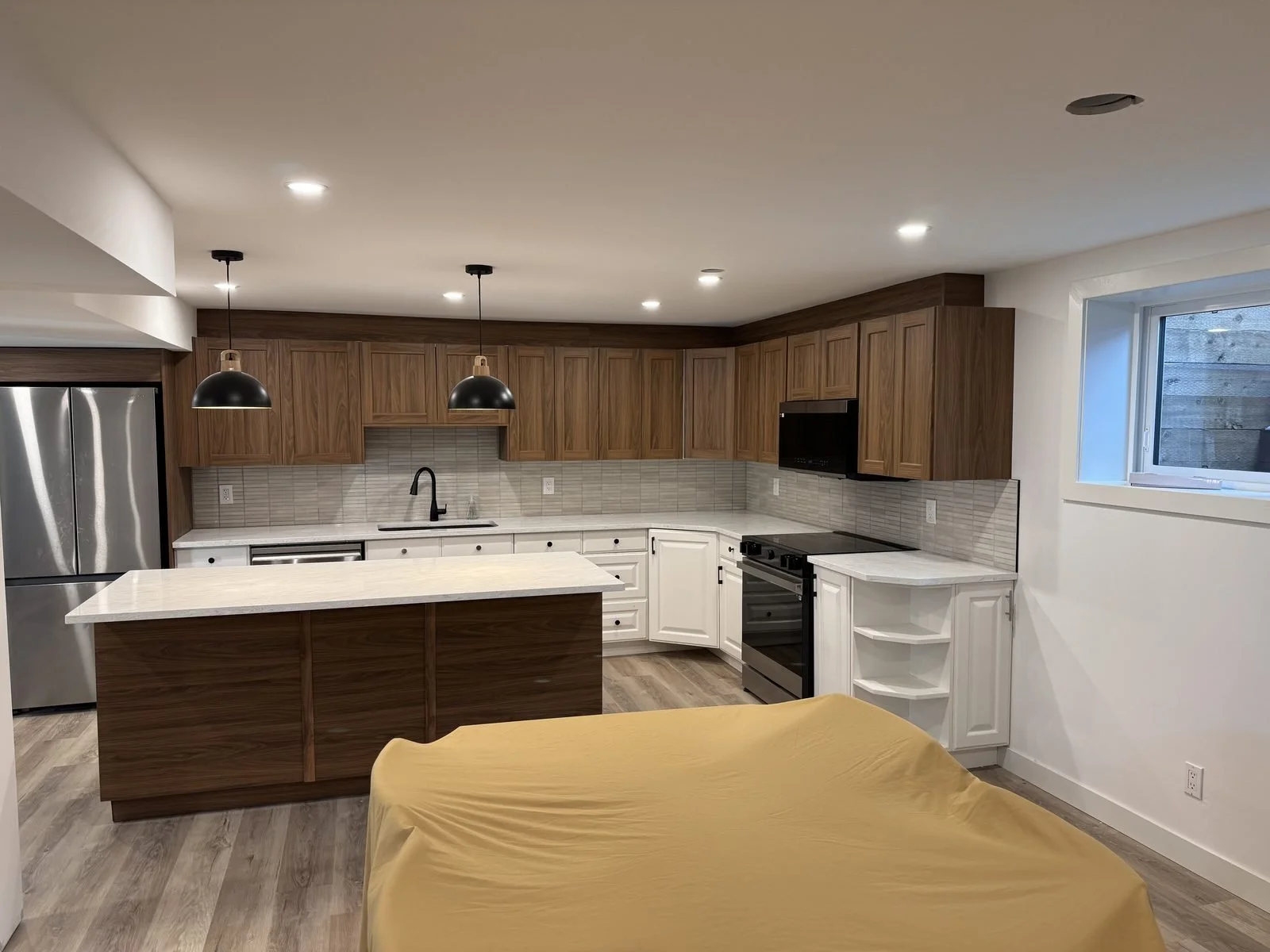 Finished legal basement suite kitchen in Midlake, Calgary, with walnut cabinets, a white island, pendant lights and pot lights wired by Koop Electrical