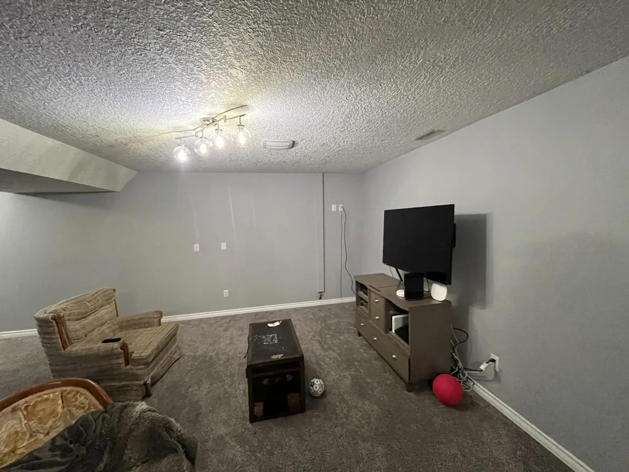 The same basement before the suite: carpeted rec room with a single track light fixture and a TV, Midlake, Calgary