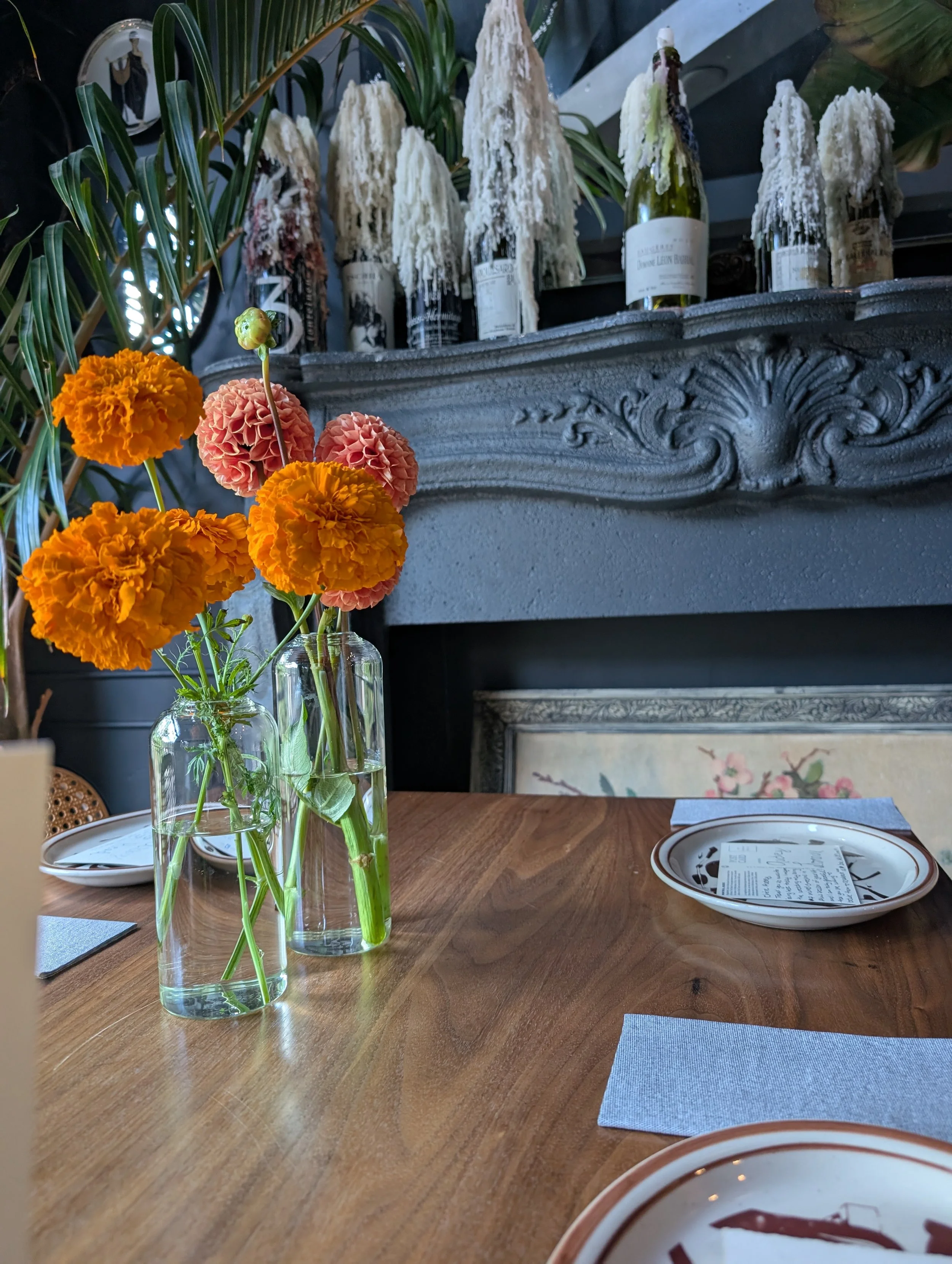 A wooden dining table with orange and pink marigold flowers in a glass vase, and place settings including a plate with handwritten note, light blue napkin, and silverware surrounded by green plants for an Ottawa wedding.
