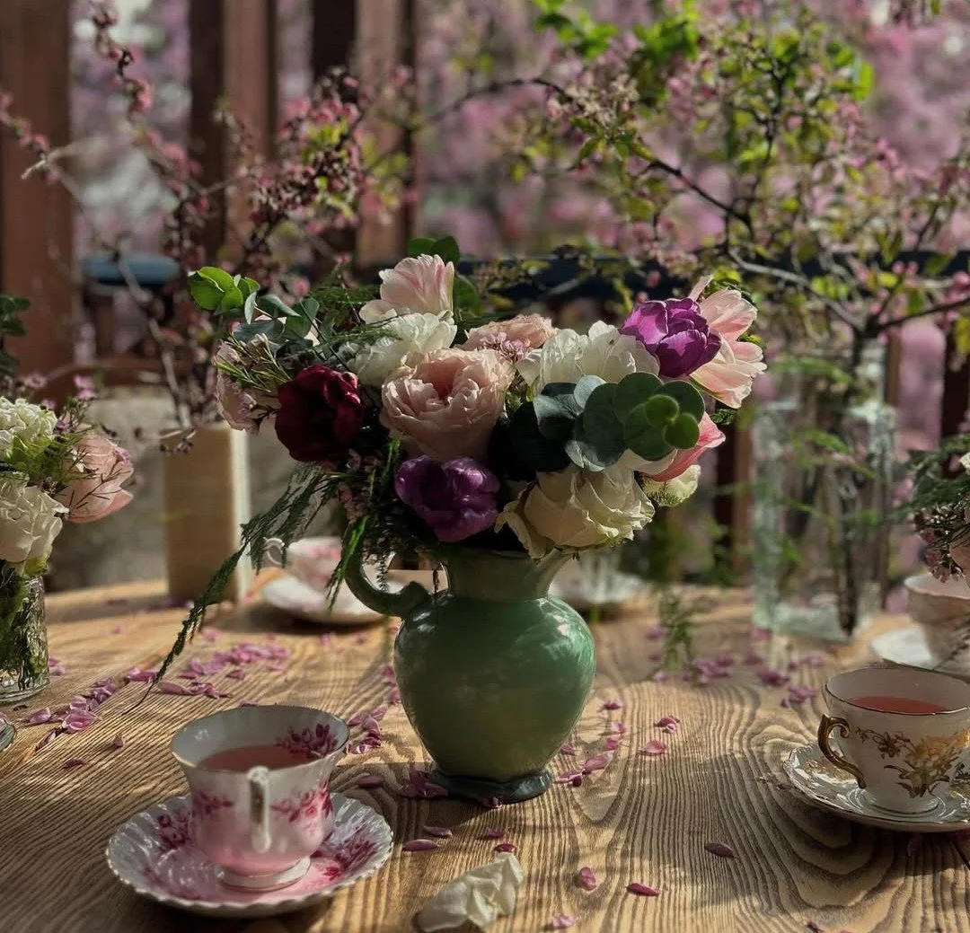 Ottawa large arrangement of pink, purple, and white flowers. Two teacups with matching saucers are on the table, and flower petals are scattered on the wooden surface. The background is filled with pink flowering branches and blossoming trees.