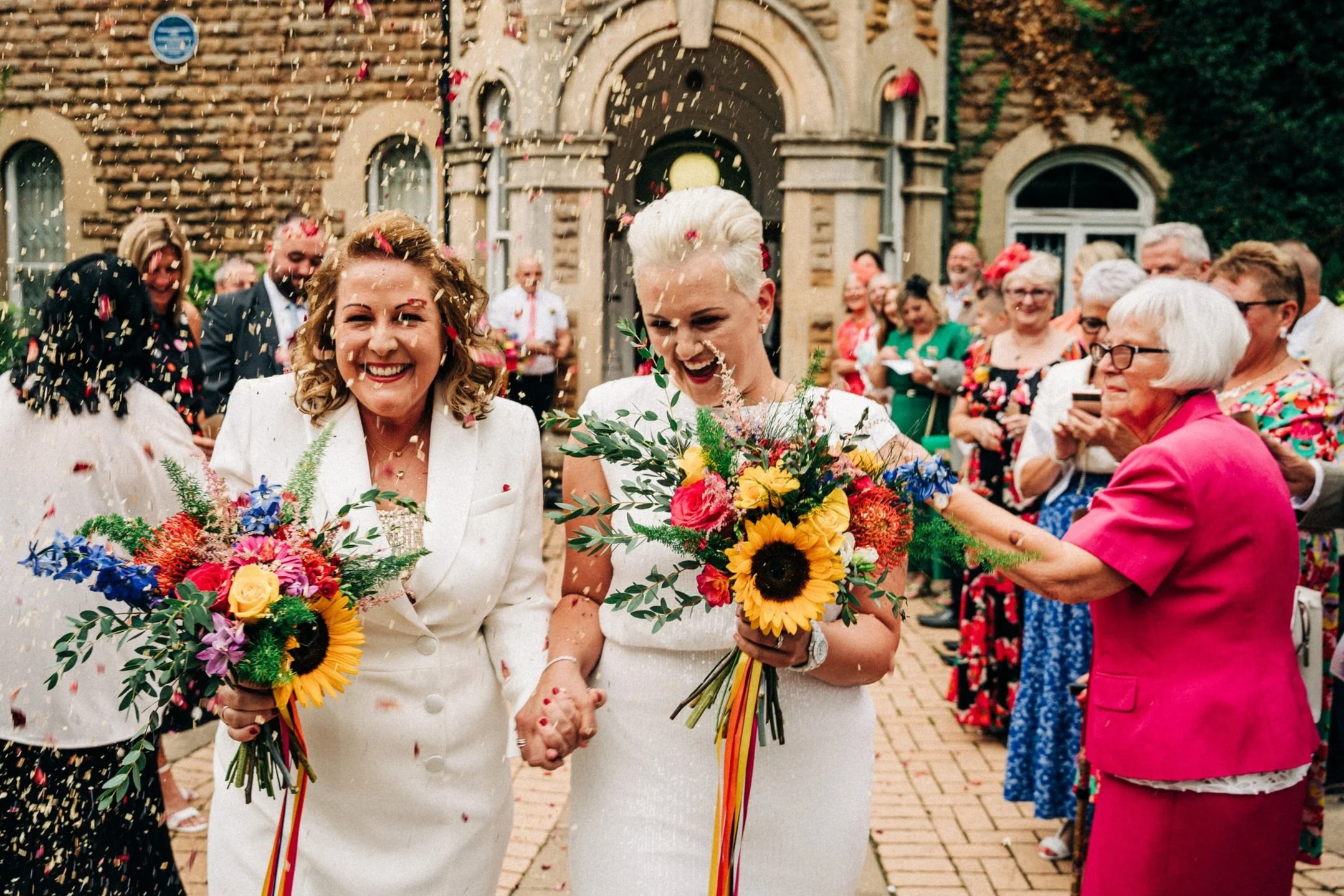 Two brides celebrating their wedding walk hand in hand surrounded by guests. They hold colorful bouquets and are showered with confetti, smiling joyfully outside a brick building.
