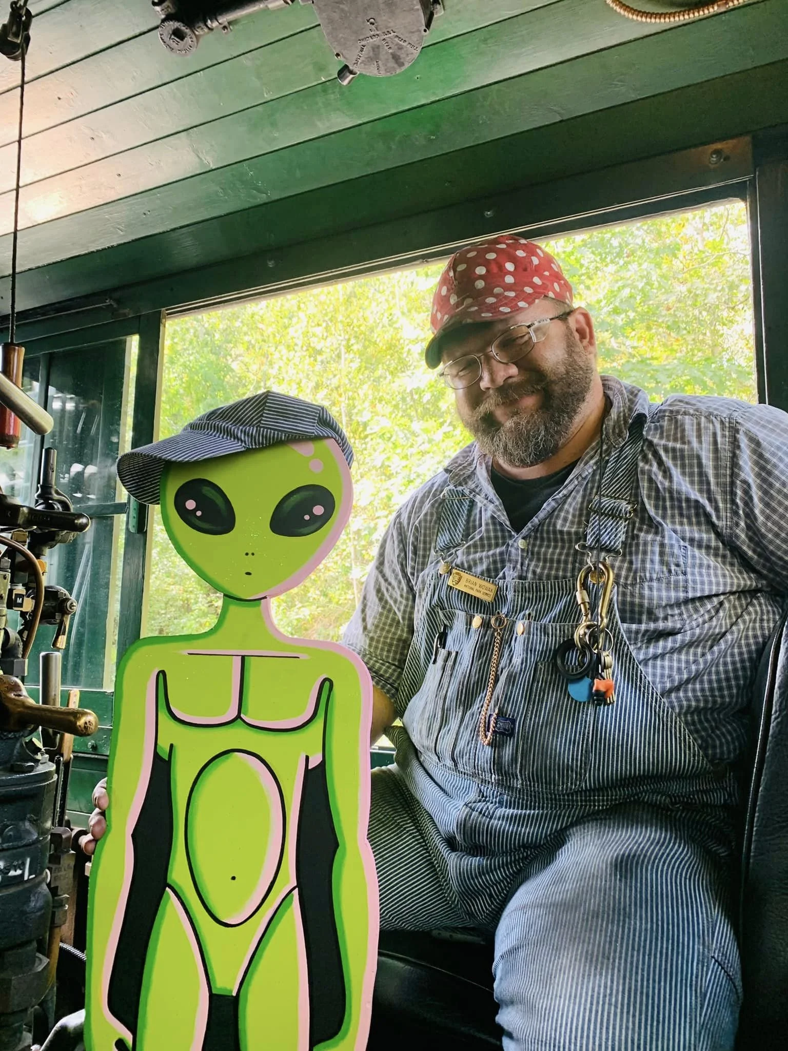 A man with glasses, a beard, wearing a red polka dot cap and an apron, standing next to a green alien-shaped cutout with a hat, inside a room with a large window and greenery outside.