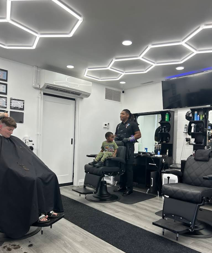 Inside Gem City Barbers in Erie, PA , a modern barbershop interior with hexagonal LED ceiling lights, black barber chairs, and a barber preparing to cut a young client's hair.