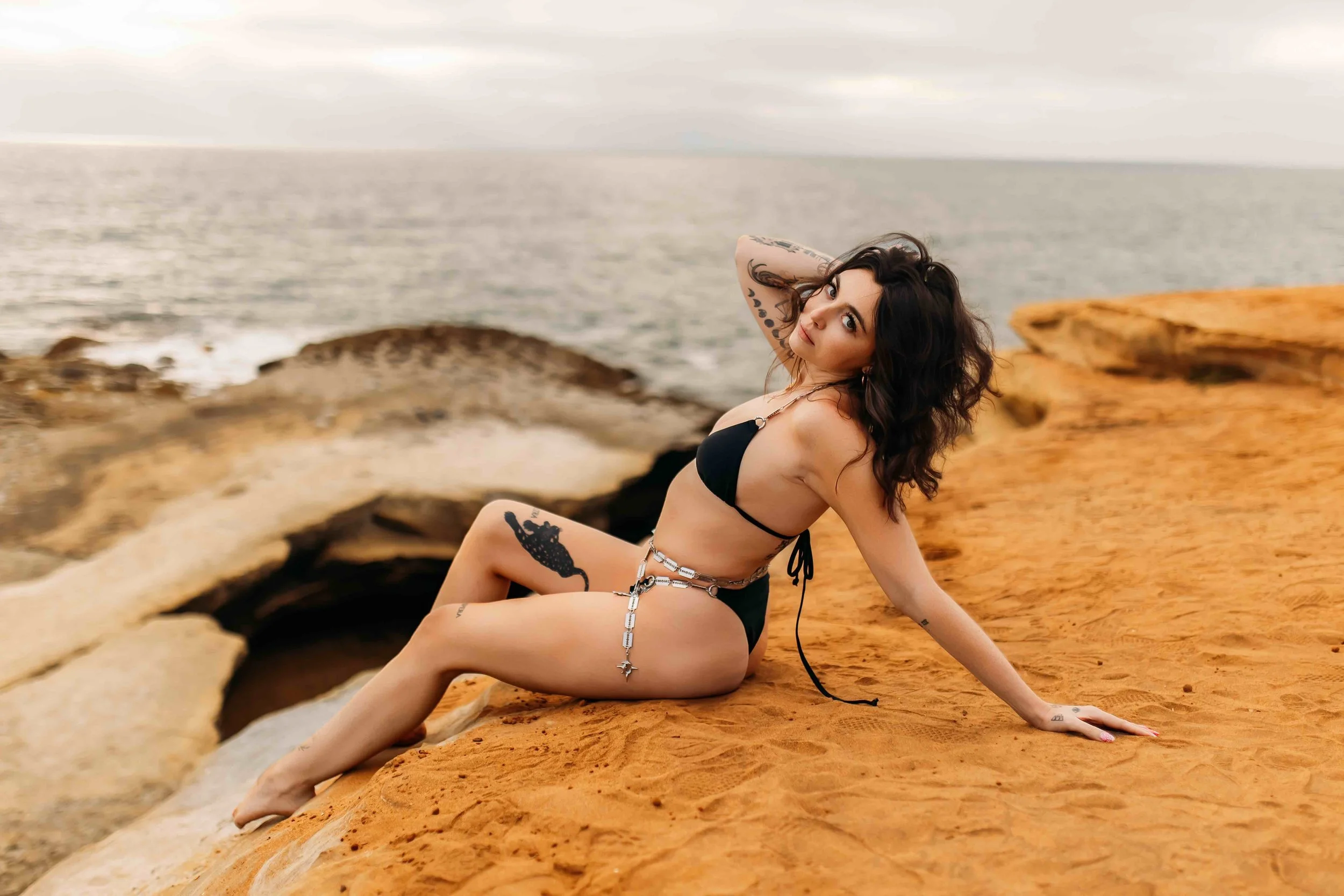 Woman in black swimsuit reclining on orange rocky surface by the ocean at sunset, with cloudy sky in background.