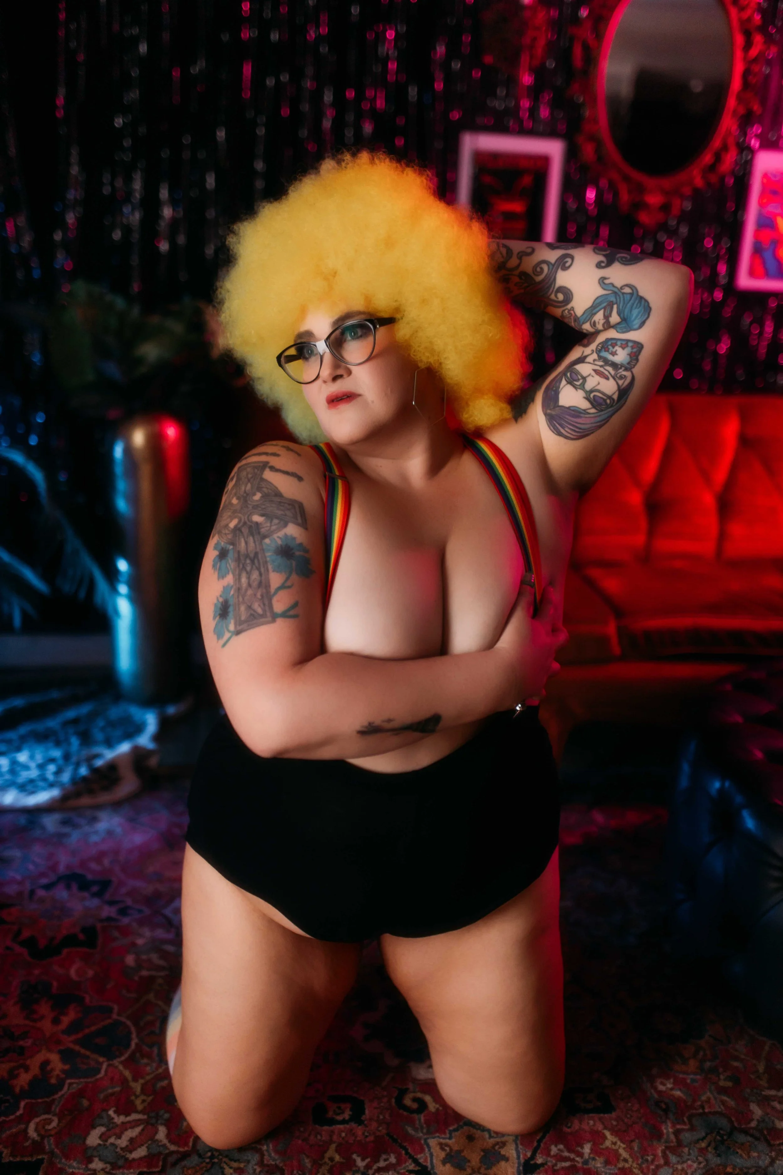 A woman with bright yellow curly hair, glasses, and multiple tattoos, wearing a rainbow strap top and black shorts, kneeling on a patterned rug in a colorful room with a red sofa and pink and black glittery walls.