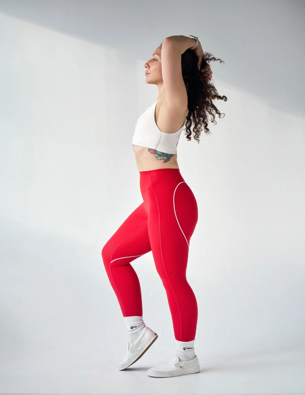 A woman with curly hair standing against a plain light gray background, wearing a white sports bra, bright red athletic leggings, white sneakers, and white socks, with her hands behind her head.