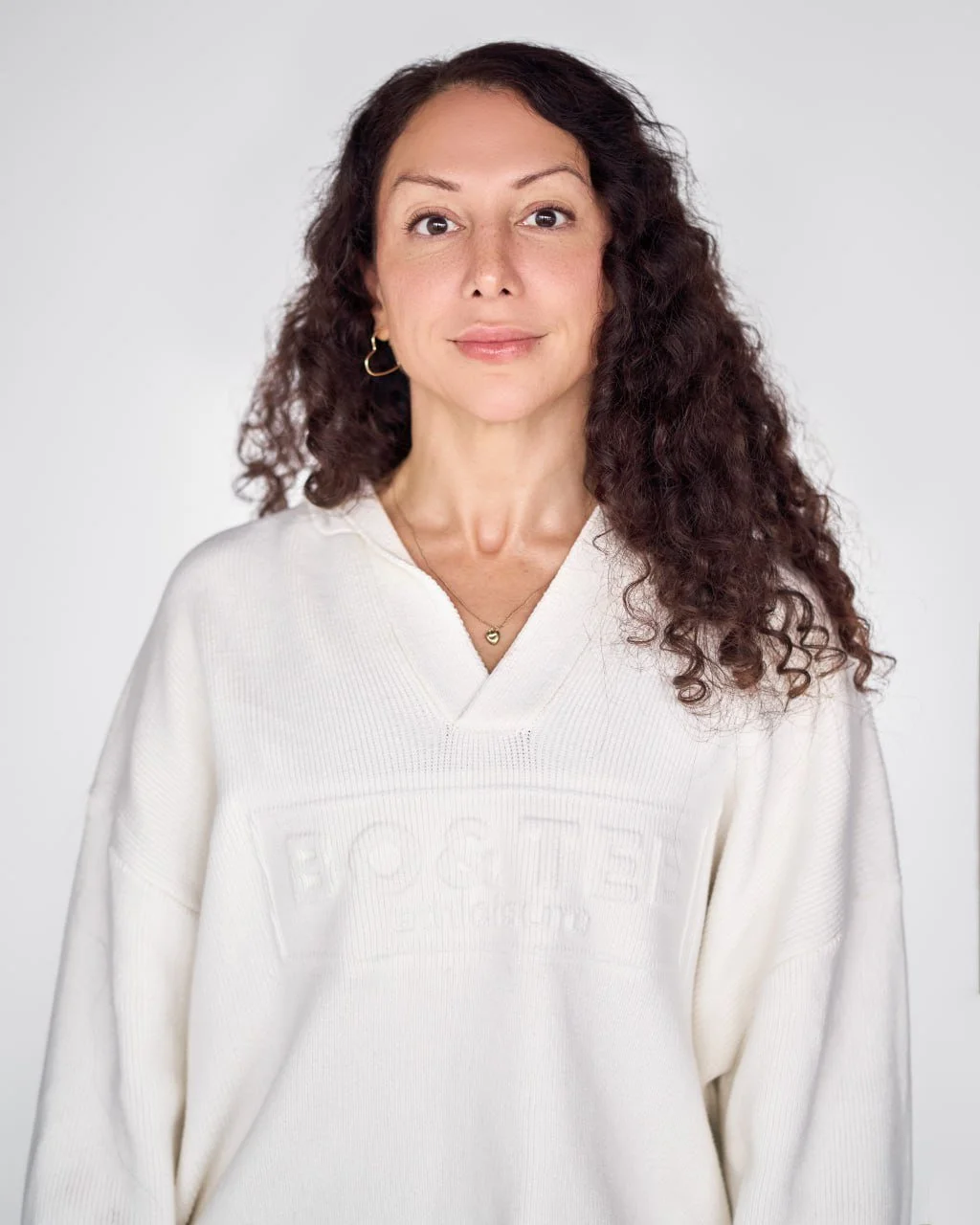 A woman with long, curly brown hair, wearing a white sweatshirt and a heart-shaped necklace, standing against a plain light grey background.