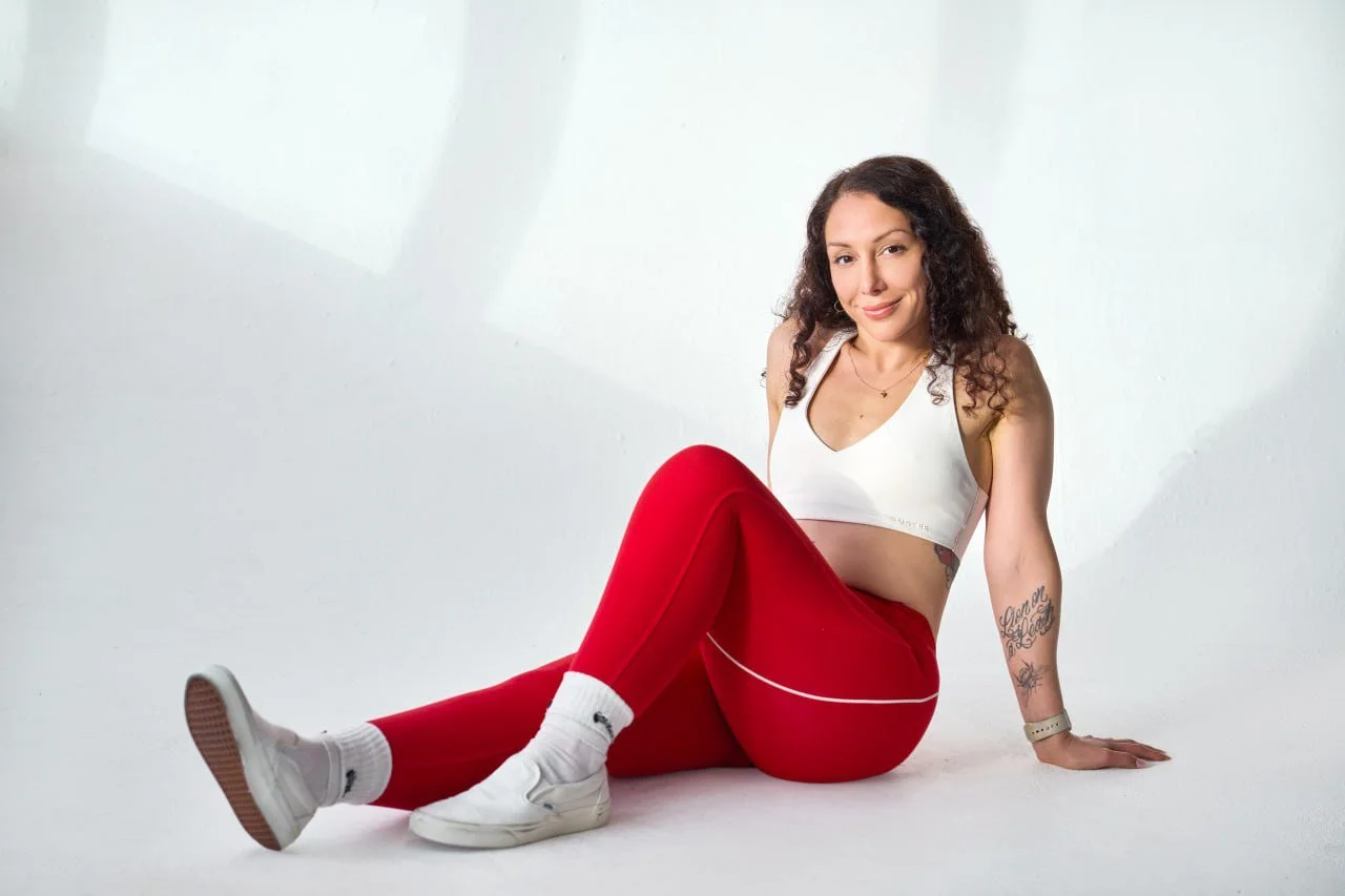 A woman with curly hair sitting on the floor against a white background, wearing a white sports bra, red leggings, white sneakers, and white socks, smiling at the camera.