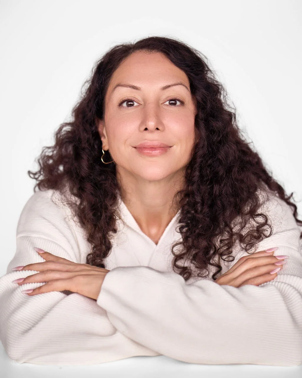 Close-up portrait of a woman with dark, curly hair wearing a white sweater, smiling softly, with her arms crossed on a white background.