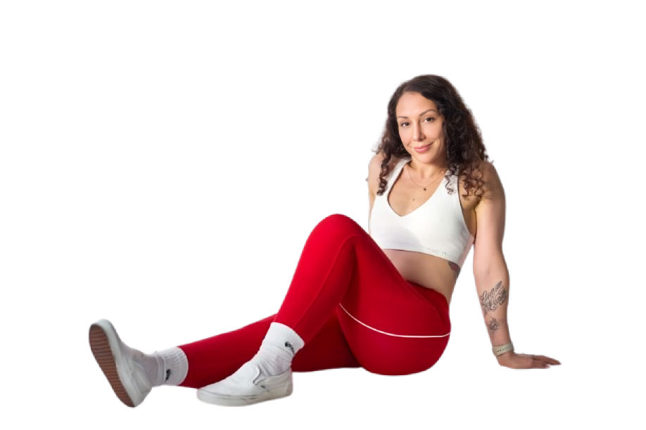 Young woman sitting on the floor with one knee up, wearing red athletic pants, white sneakers, a white sports bra, and smiling at the camera.