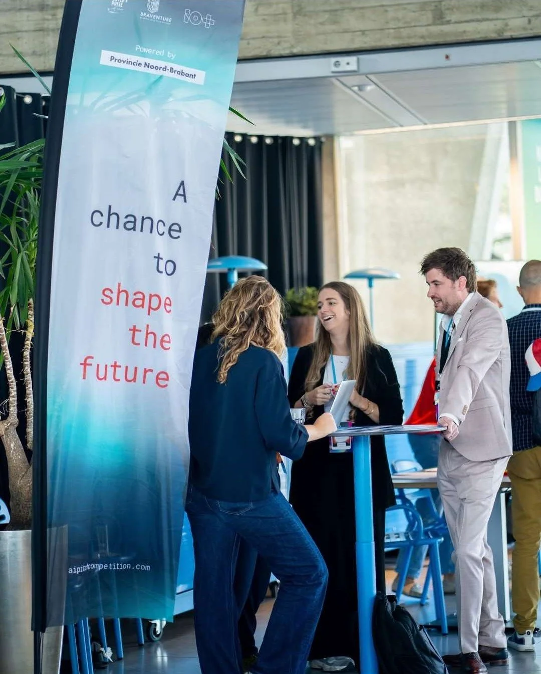 People at an AI Pitch Competition workshop engaging in conversation, with a vertical banner displaying the text 'A chance to shape the future' and mentioning 'Powered by Provincie Noord-Brabant'.