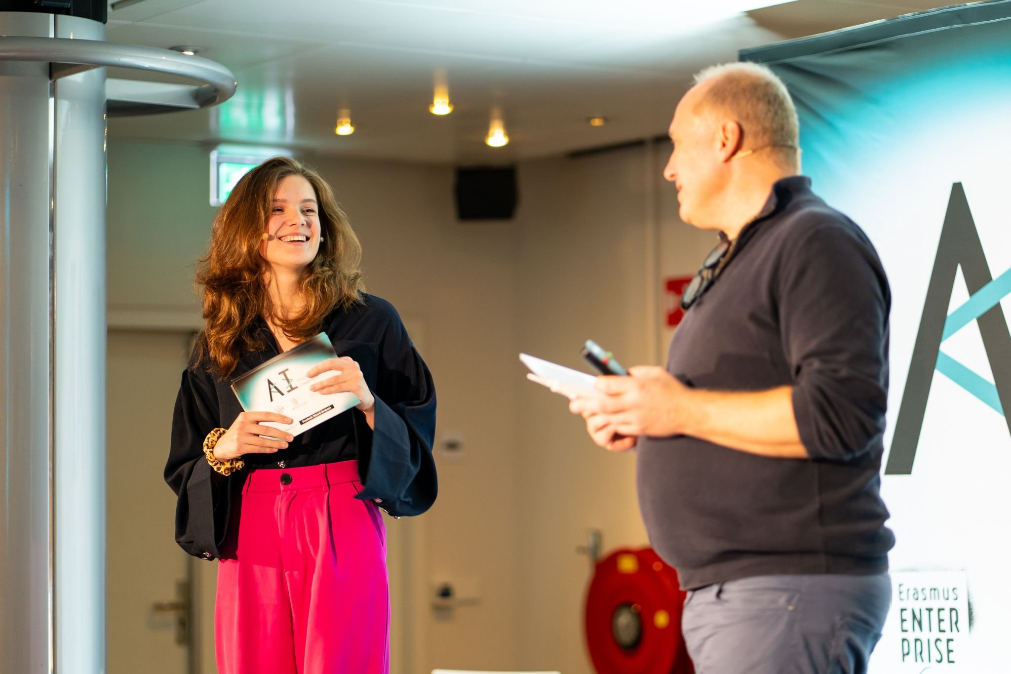 A woman with long brown hair smiling and holding a speaker card with AI Pitch Competition logo, engaging in conversation with a man who is holding a notepad and pen, standing on a stage at the finale of AI Pitch Competition.