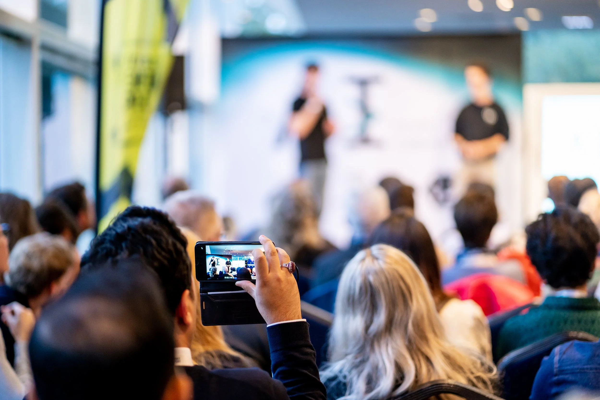 A person in the foreground is holding a smartphone, recording or taking a photo of an AI Pitch Competition finalists' pitch among a large audience.