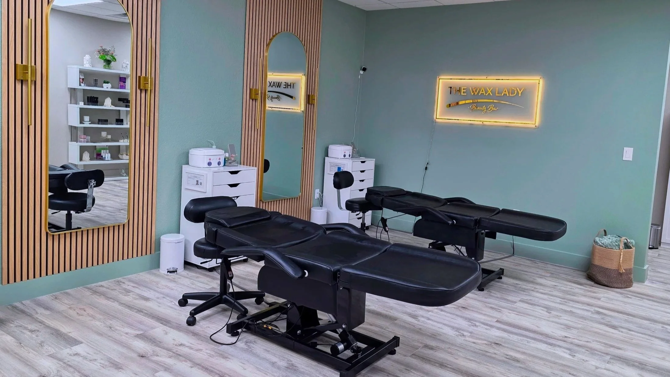 Interior of a beauty treatment room with two black adjustable massage or treatment chairs, a mirror on the wall, and a neon sign reading 'The Wax Lady' in script font. There are small white cabinets with equipment and supplies, and a basket with towels on the floor.