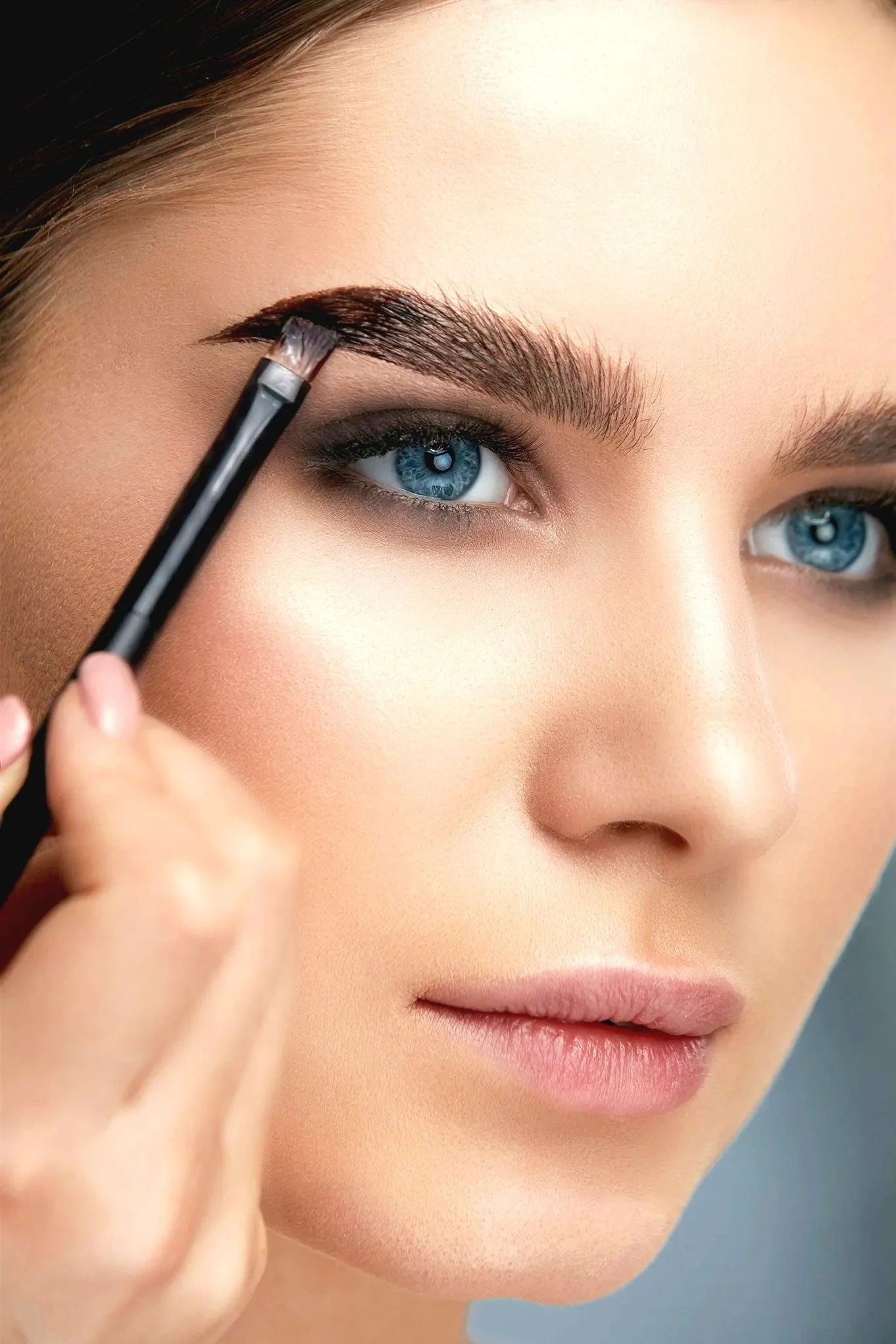A woman with blue eyes applying tint to her eyebrows with a brush.