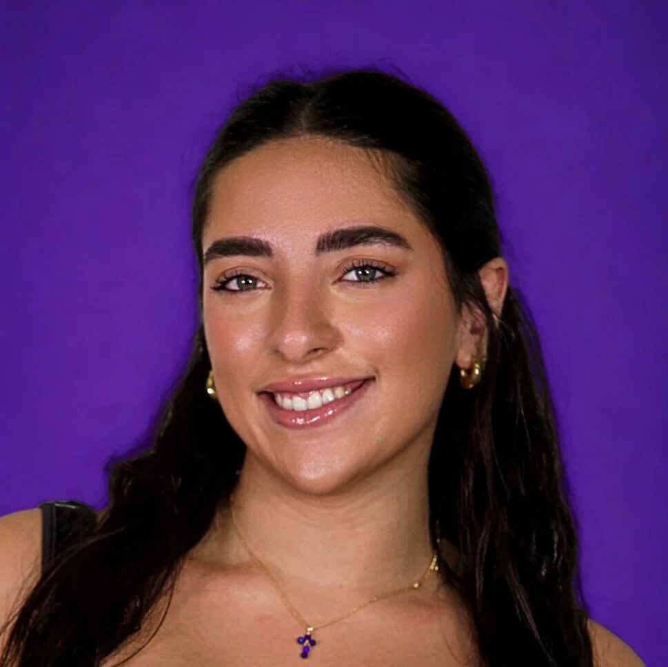 Close-up portrait of a smiling young woman with dark brown hair, green eyes, wearing gold earrings and a gold necklace with a small blue pendant, against a purple background.