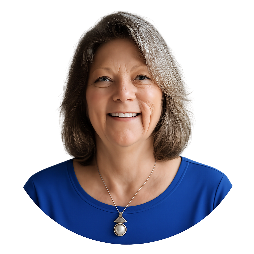 A smiling older woman with shoulder-length gray hair, wearing a blue floral top and a necklace with large pearl and triangular pendant. She is sitting indoors near a window.