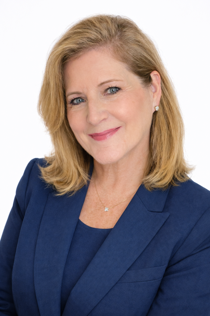Professional portrait of a woman with blonde hair, smiling, wearing pearl earrings, a white shirt, and a black blazer, against a grey background.