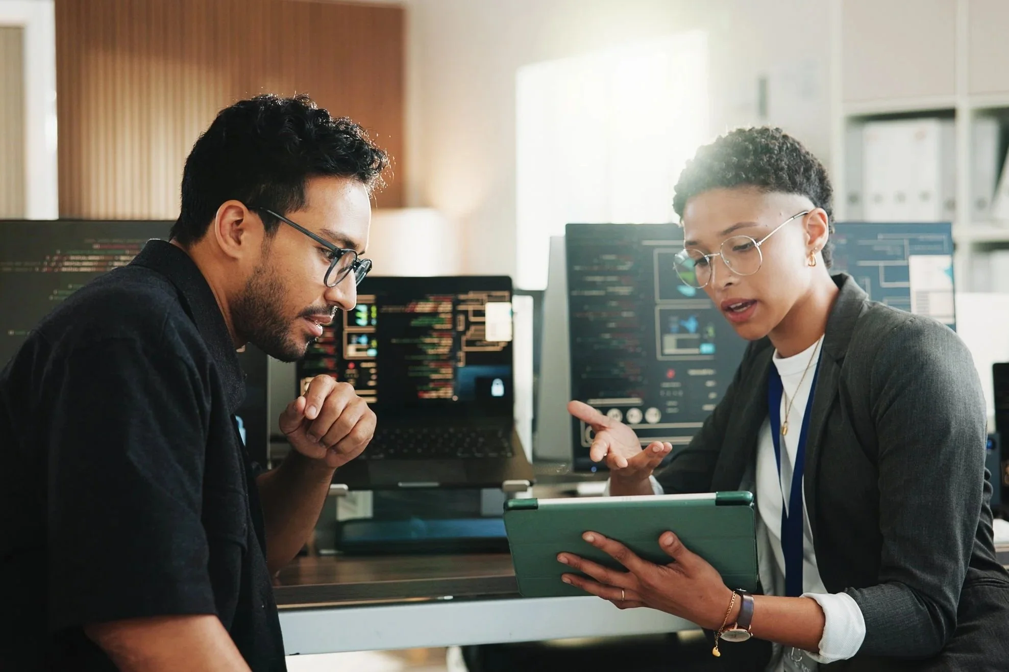 Two colleagues review a tablet together in an office lined with monitors