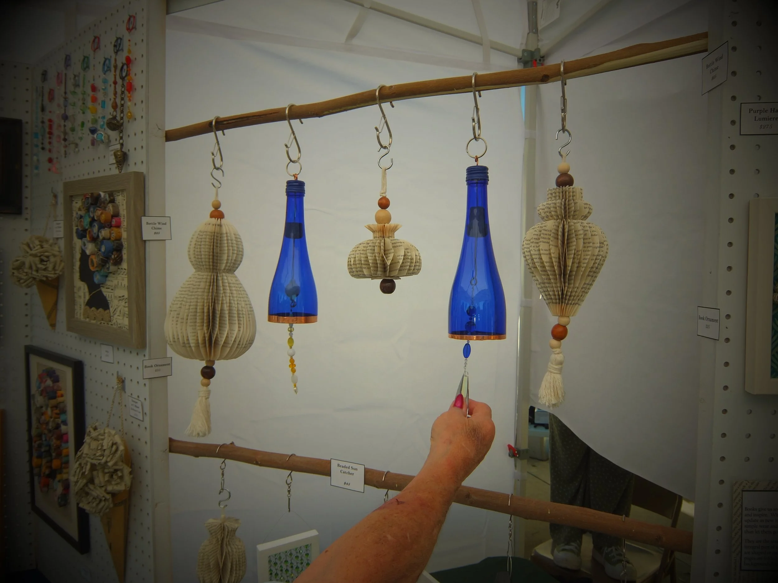 Hand holding a beaded sun catcher with blue bottles and paper origami decorations hanging from a wooden rod at a craft fair.