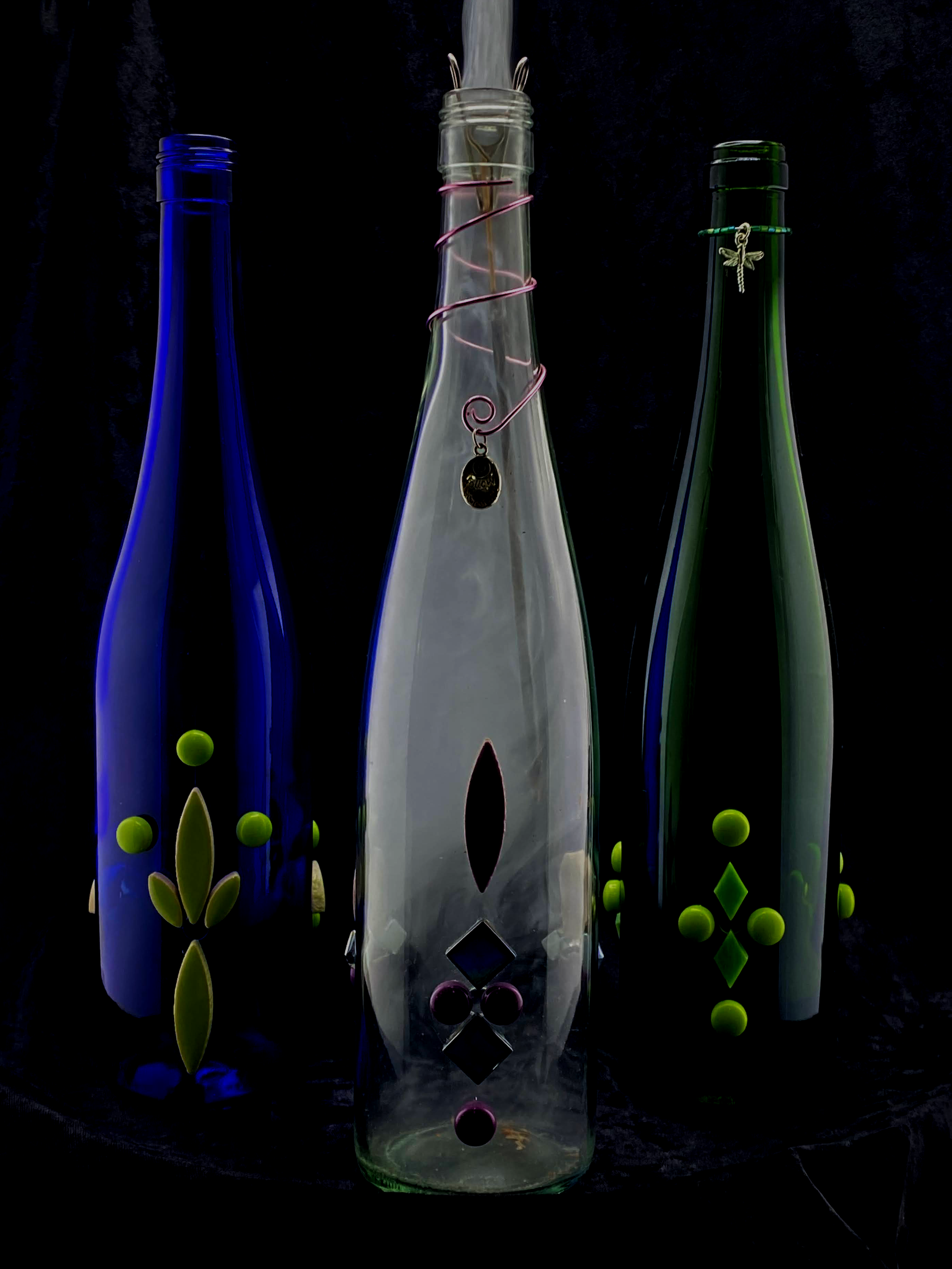 Colorful glass bottle art with geometric and organic shapes, including a leaf and a pin with a dragonfly charm, set against a dark background.
