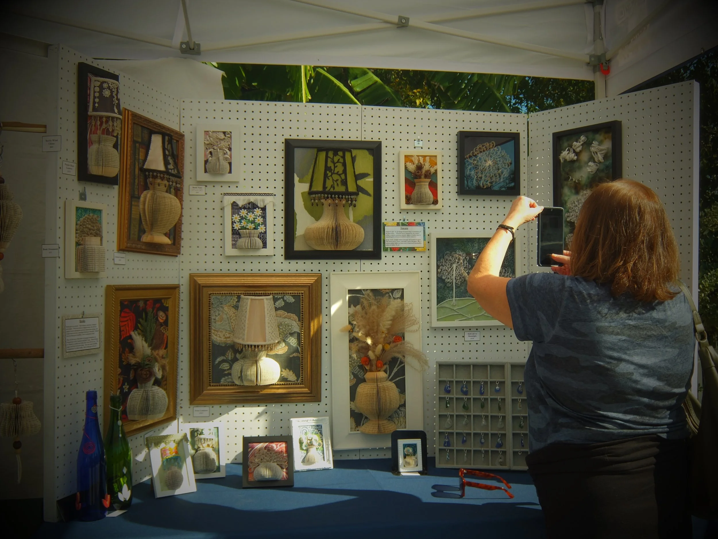 Art display booth featuring framed and unframed fiber art and woven baskets at an outdoor craft fair, with a person taking a photo.