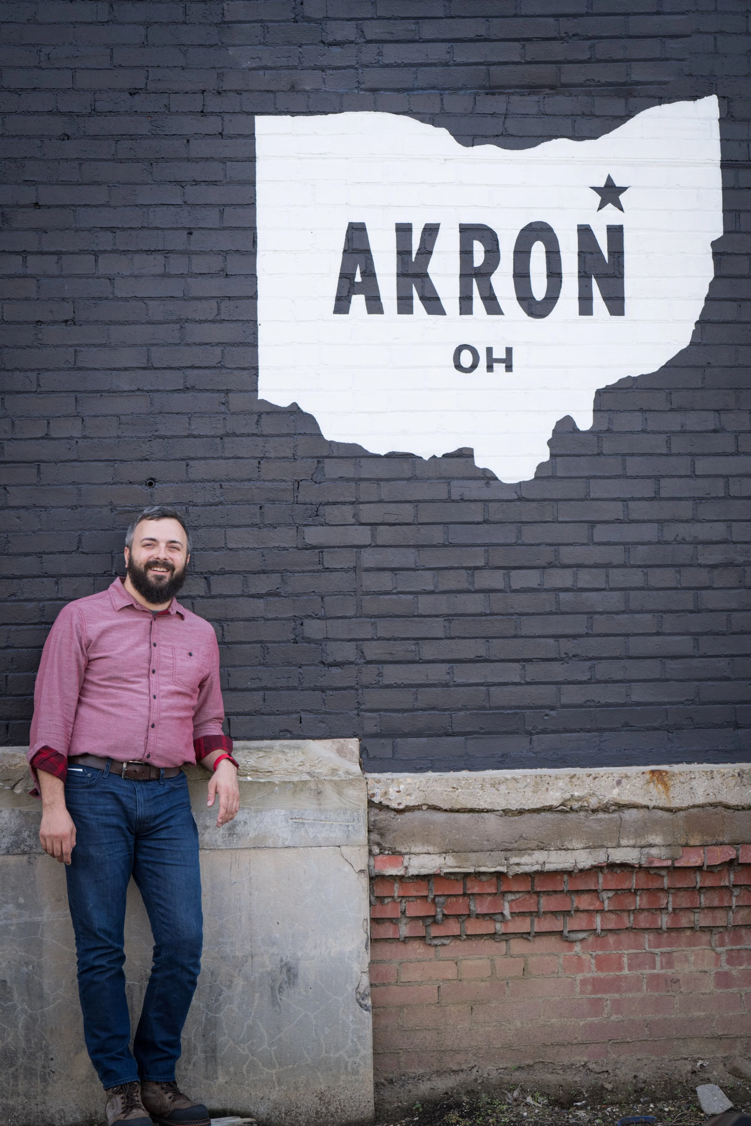 A man with a beard and a pink shirt standing in front of a brick wall with a large painted outline of the state of Ohio and the word 'AKRON' inside it.